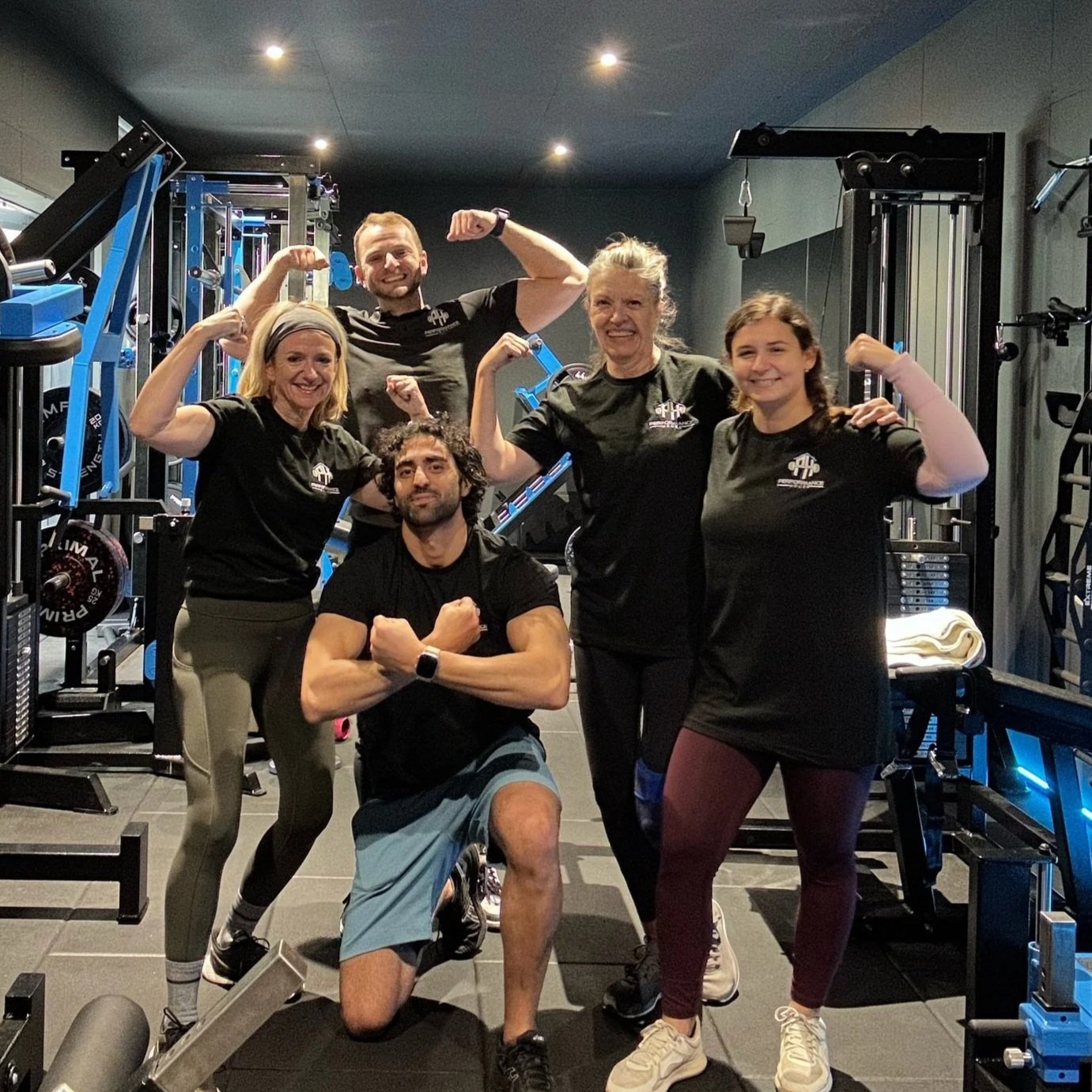 Group of five people at the gym, flexing their muscles and smiling, with gym equipment in the background.