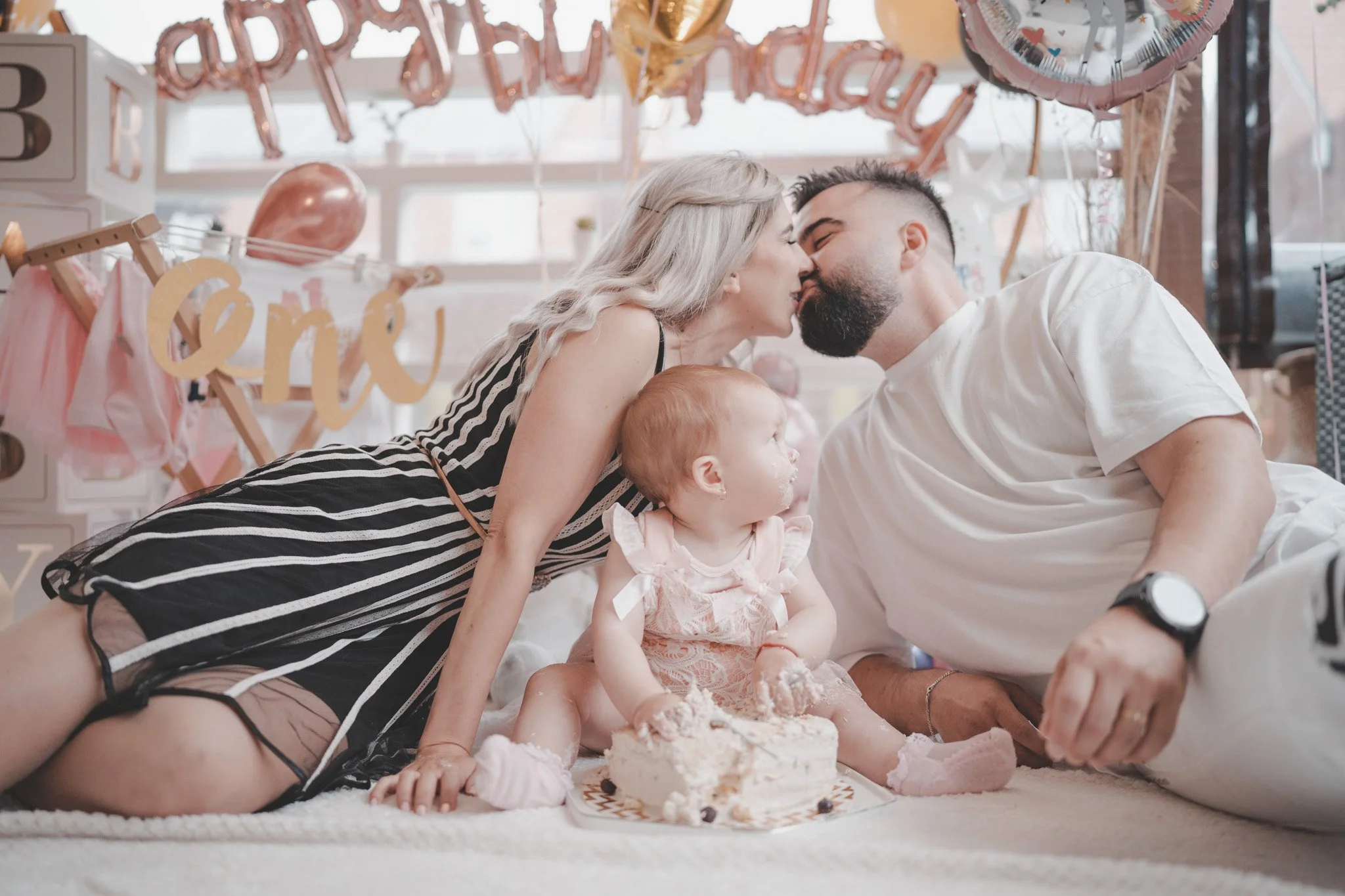 Parents and their one year old daughter sitting on the floor and giving each other a kiss at a smash the cake party in Coventry