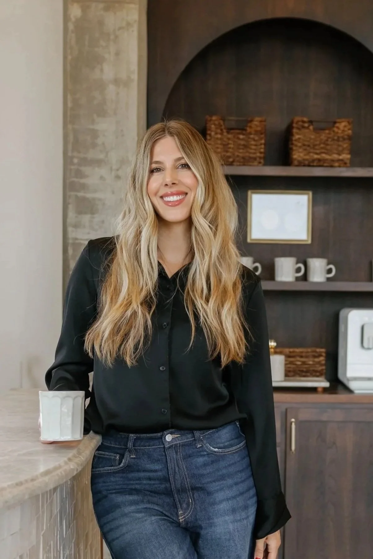 A woman with long, wavy blonde hair and a bright smile, standing in a modern kitchen with a coffee mug in her left hand.