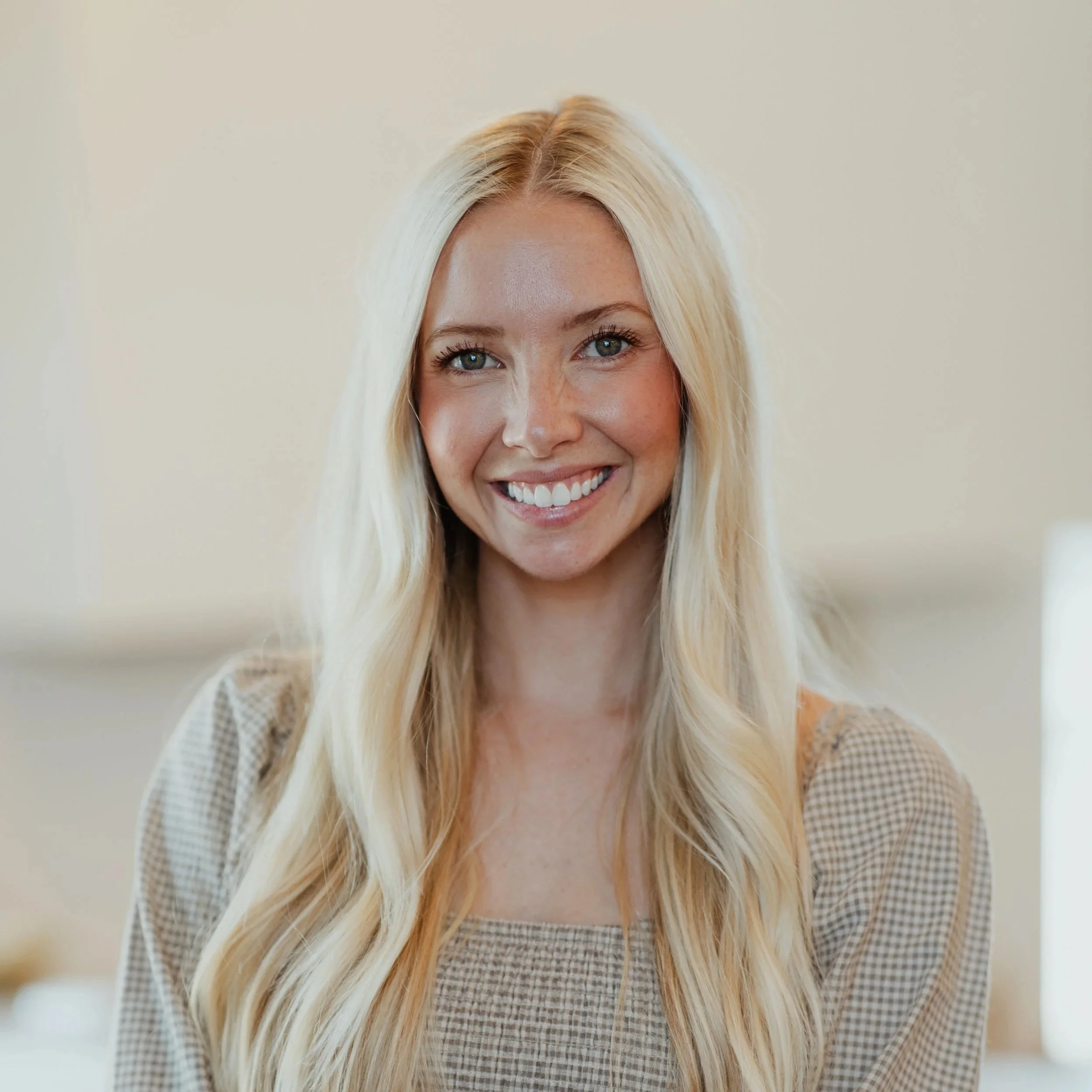 Portrait of a smiling woman with long blonde hair and blue eyes, wearing a beige checkered top, indoors with a neutral background.