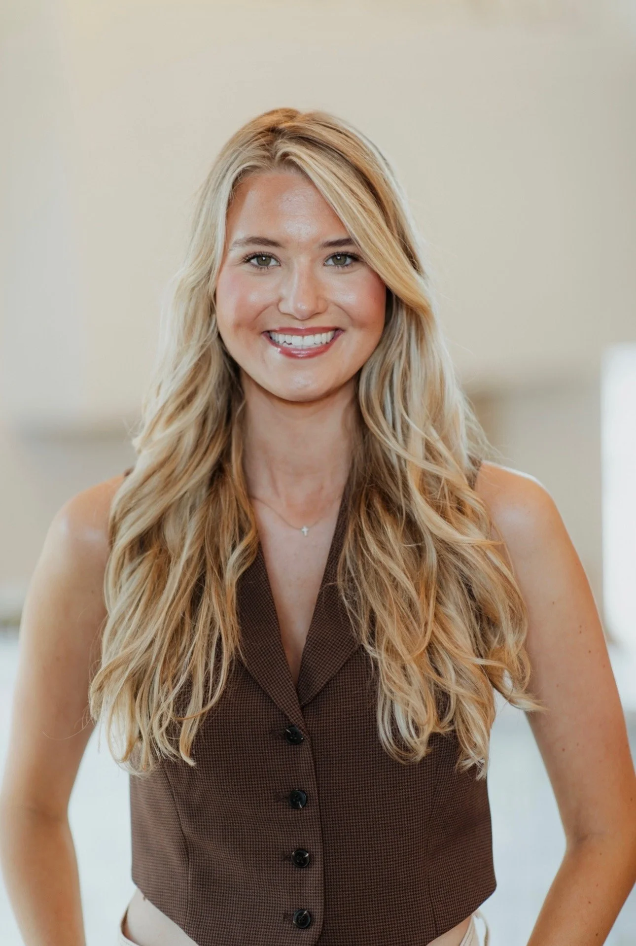 A young woman with long wavy blonde hair smiling, wearing a sleeveless white button-up top and a delicate cross necklace, against a plain white background.