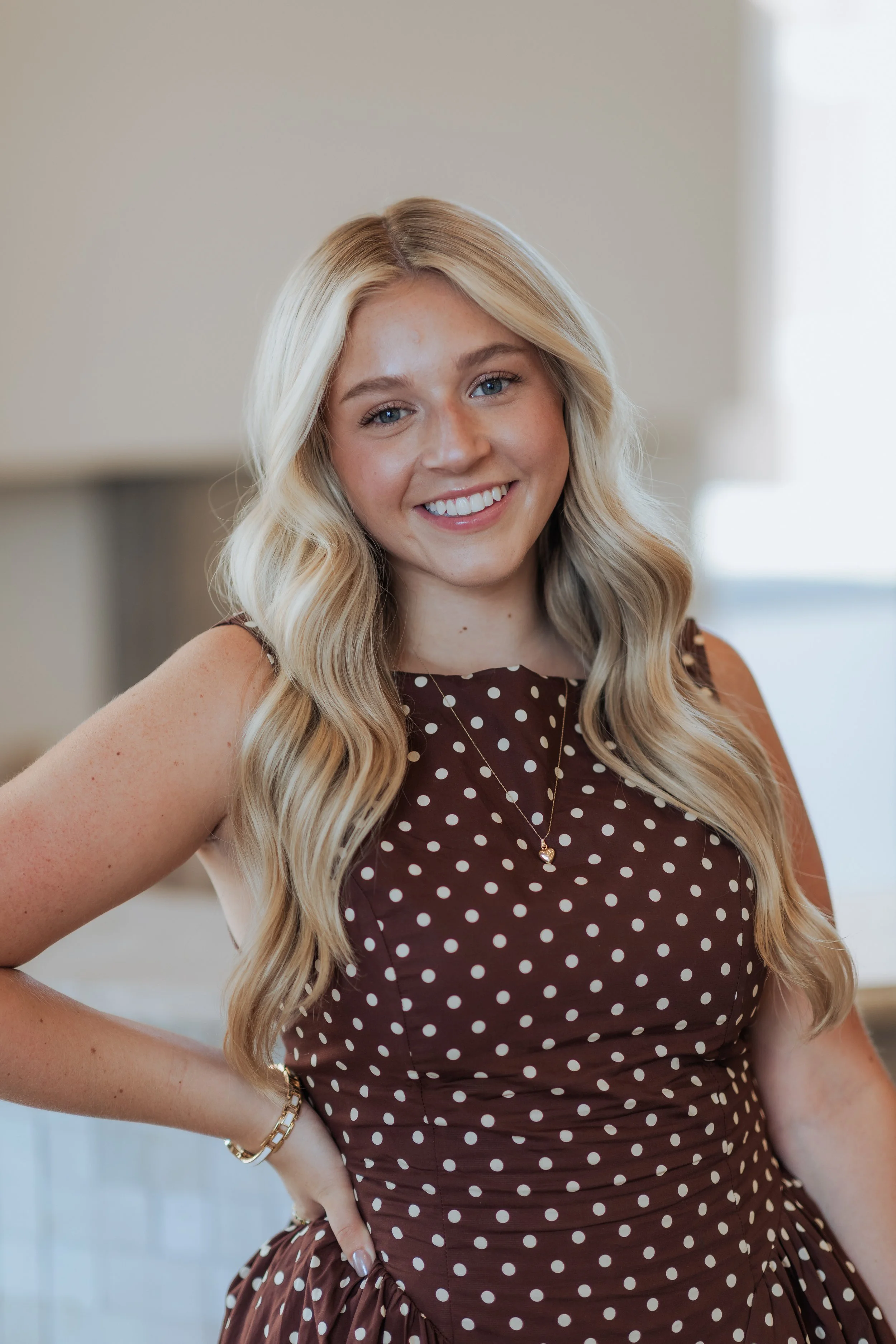 A smiling blonde woman with wavy hair wearing a black top with white accents, standing against a white background.
