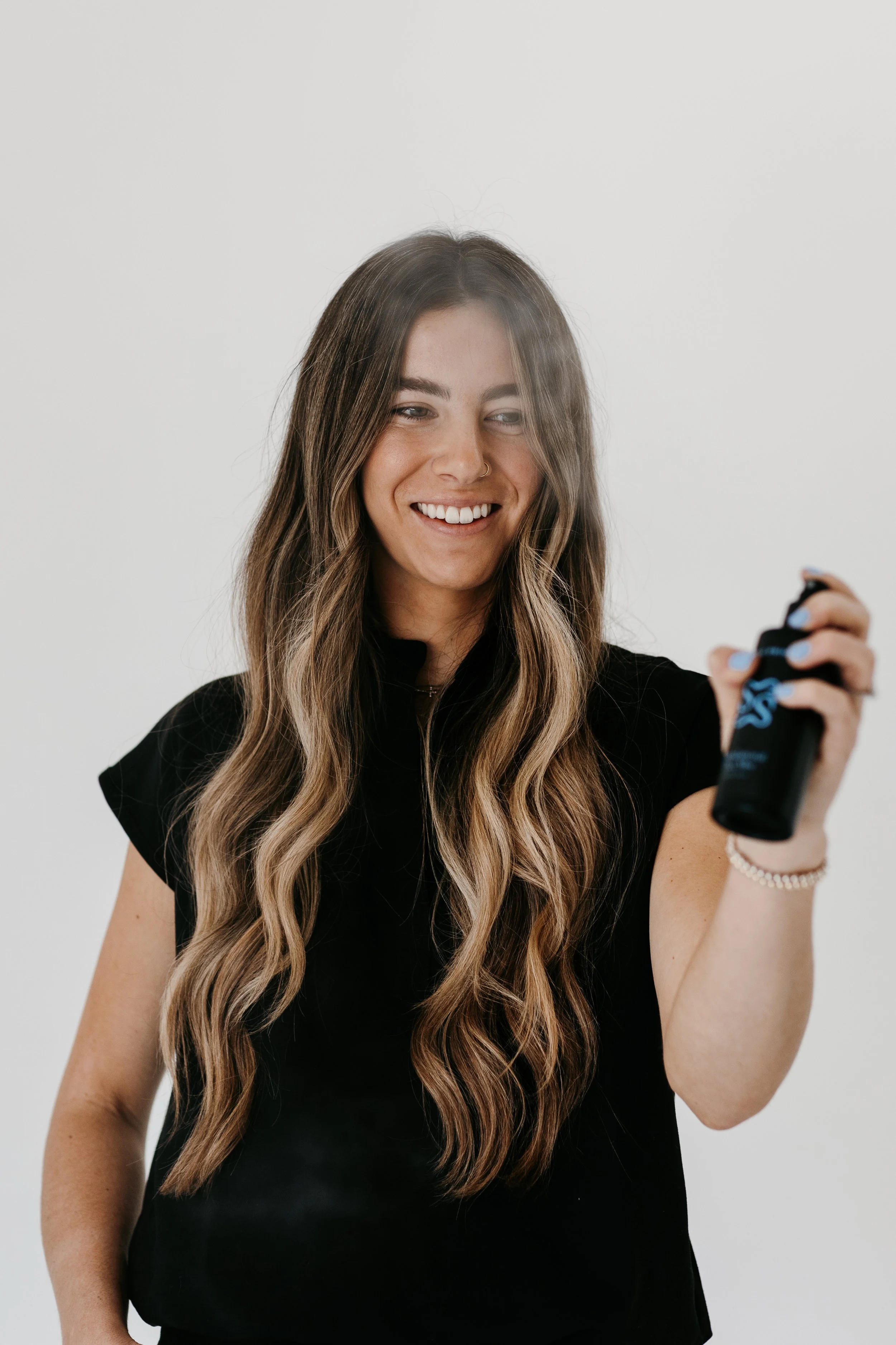 A young woman with long wavy brown hair is smiling and holding a black spray bottle in her right hand, standing against a plain white background.