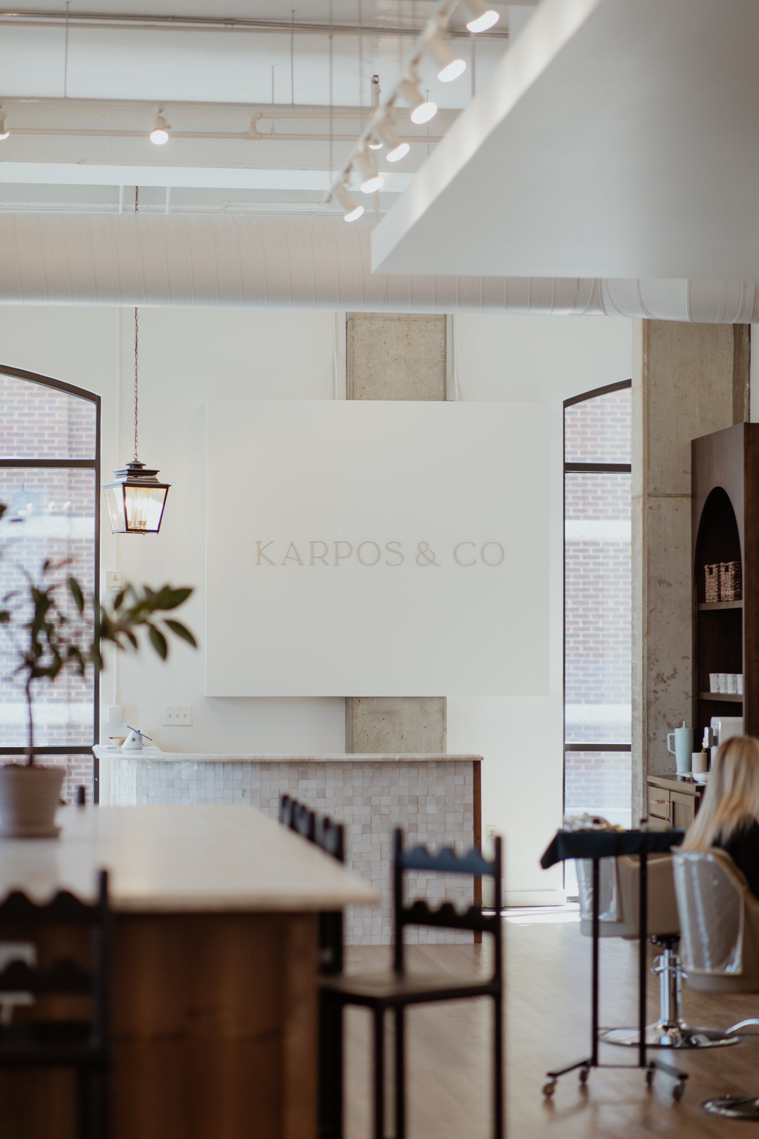 Interior of a modern hair salon with a white wall displaying 'KARPOS & CO' and natural light from large windows.