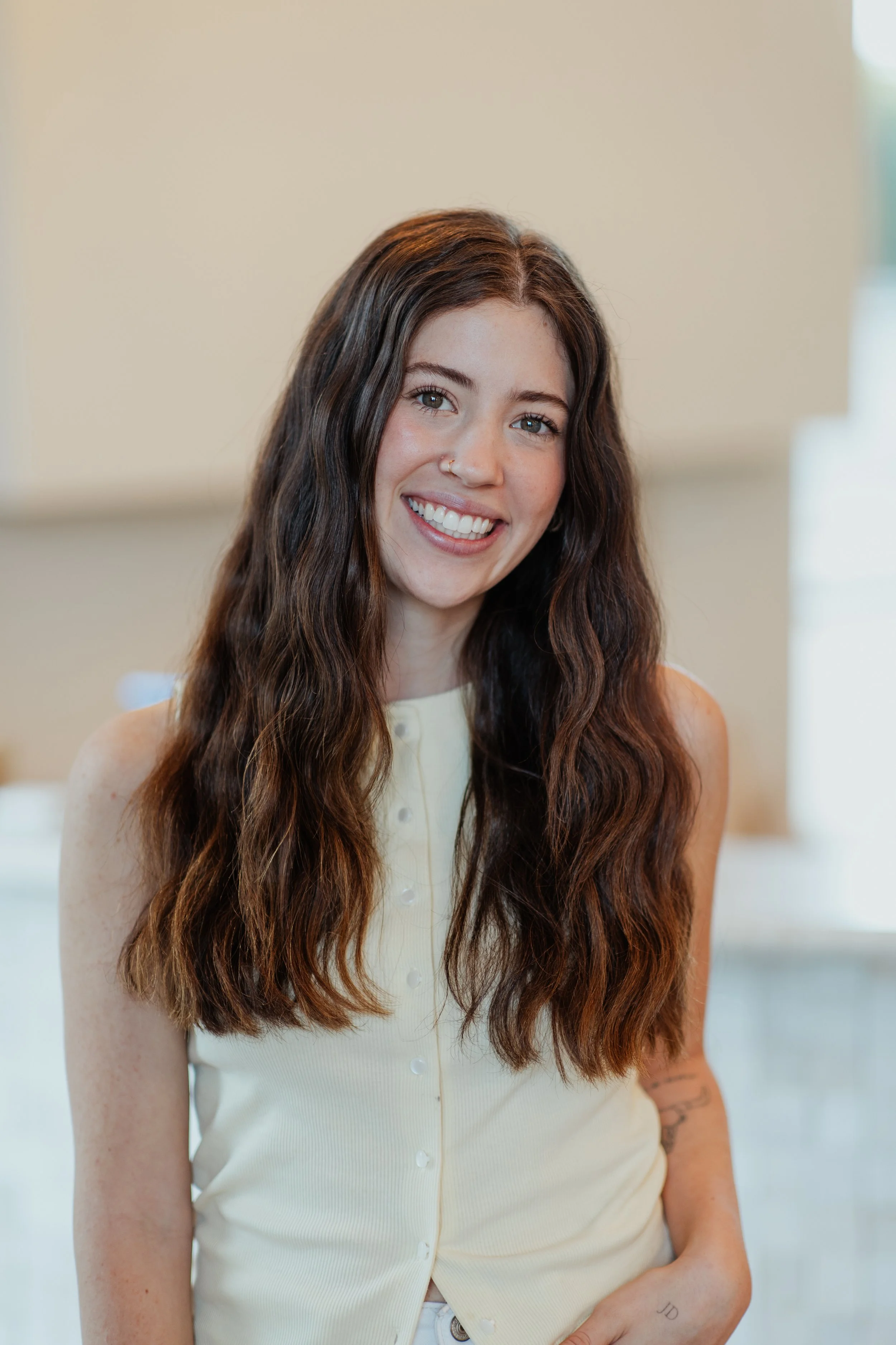 A young woman with long, wavy brown hair smiling indoors, wearing a sleeveless light yellow top and a nose ring.