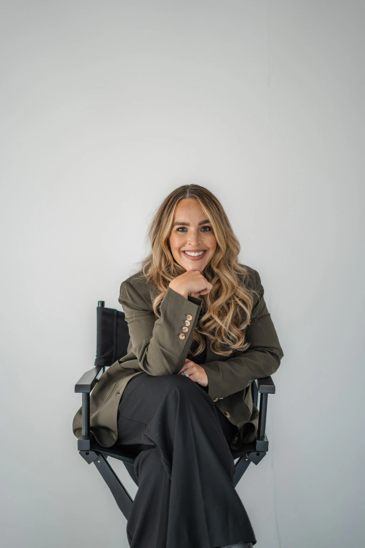 A woman with wavy blonde hair, smiling, wearing hoop earrings, a black top, a beige blazer, and a black belt, standing indoors with a lamp, a potted plant, and a framed photo in the background.
