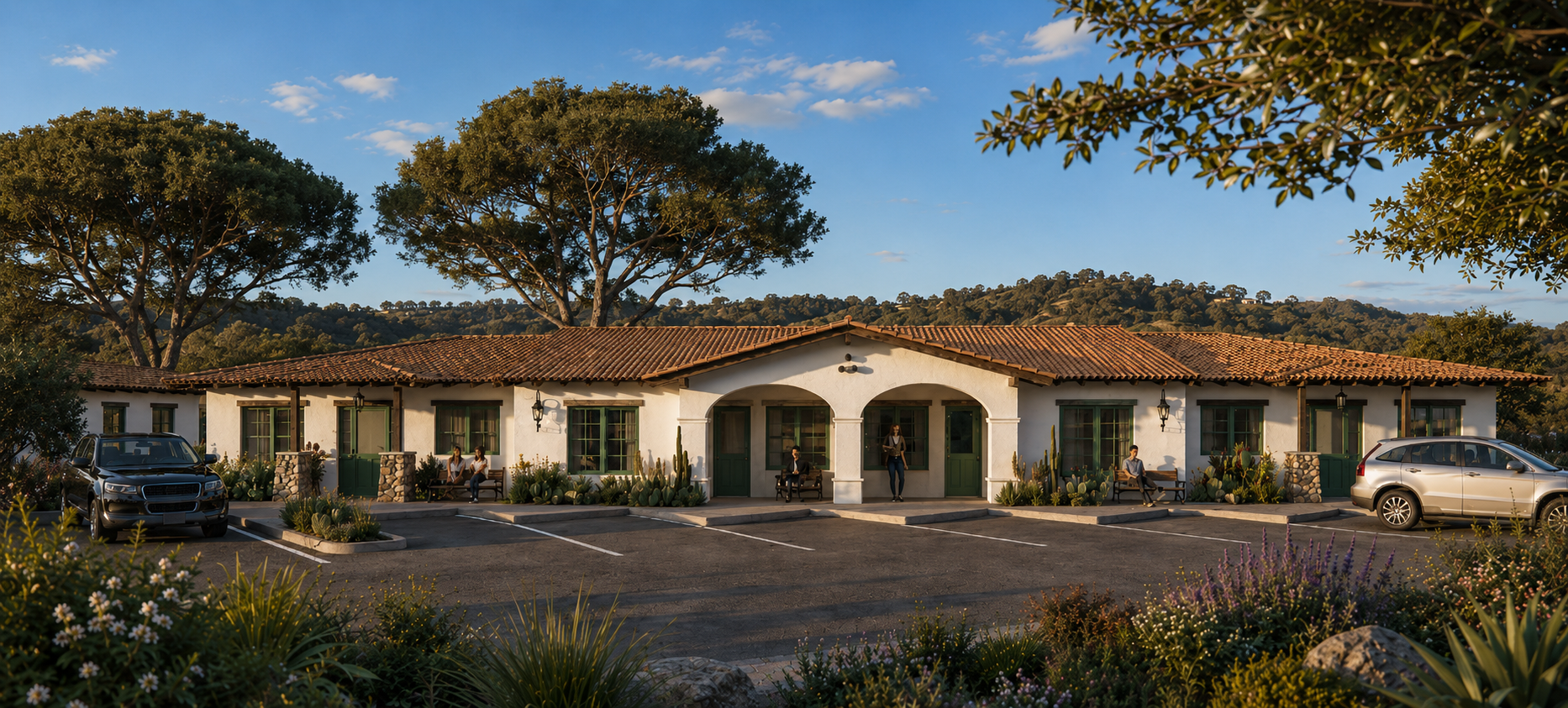 A row of white buildings with red doors and windows, and desert landscaping, including cacti and succulents, under large trees. People are sitting and standing outside, and cars are parked in front. The scene appears to be in a sunny desert environment.