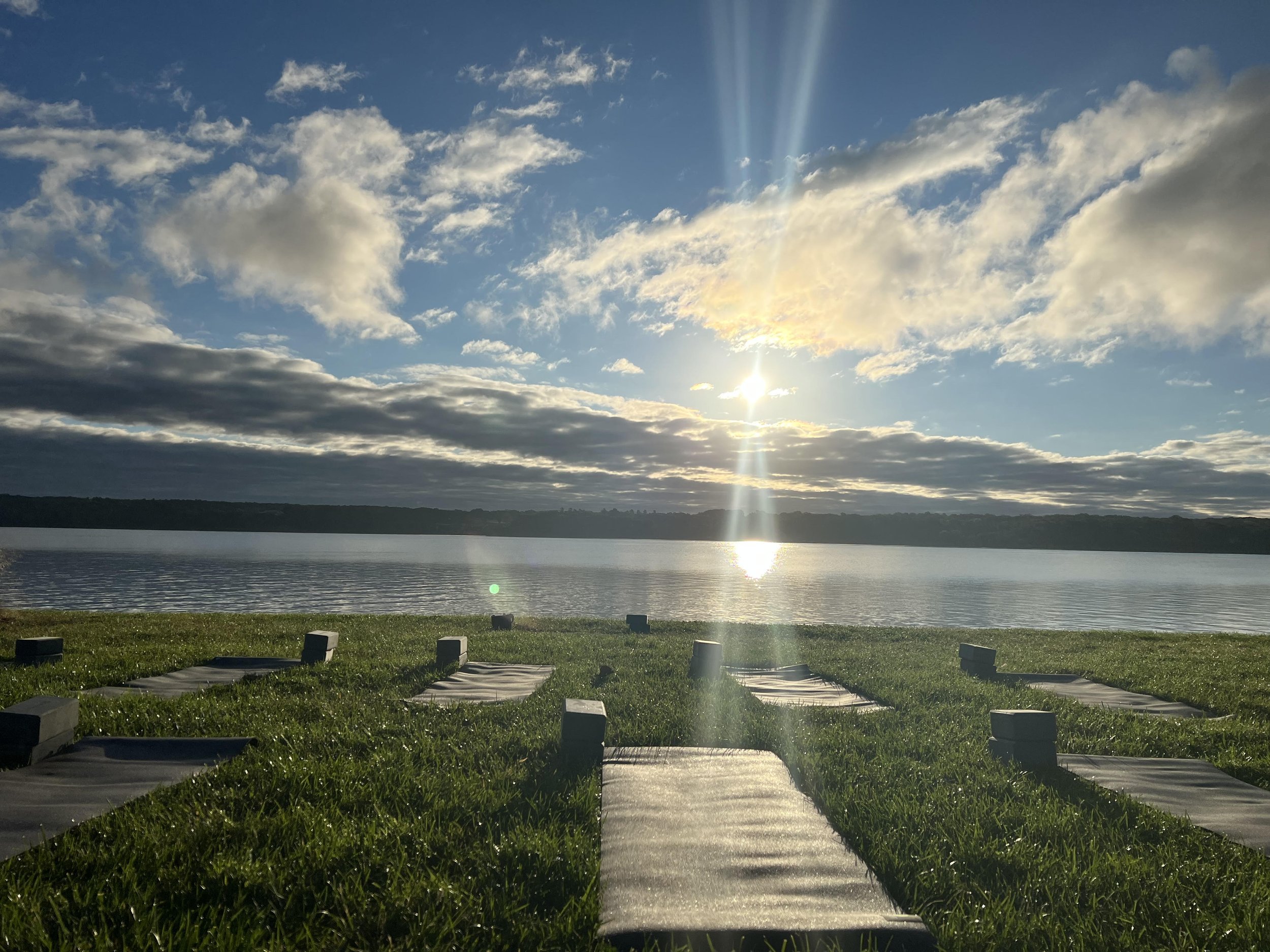 Yoga mats and blocks on grass by a lake during sunrise with clouds and sunlight reflections on water.