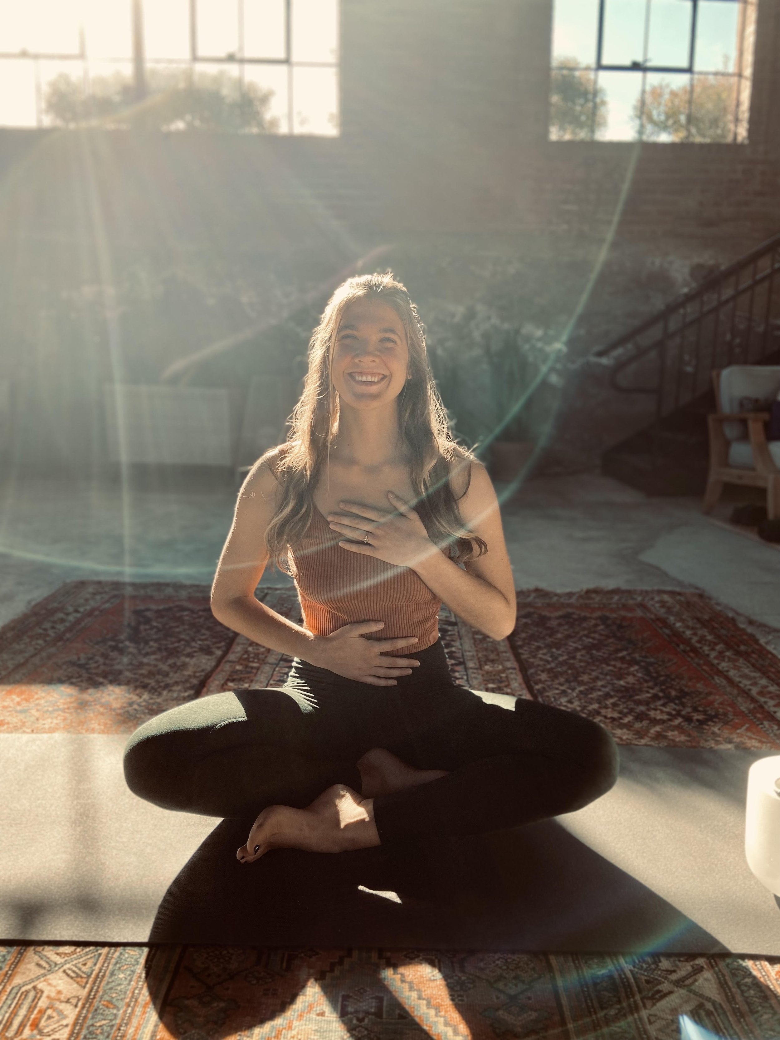 Joey-Lynn Henderson sitting cross-legged on a yoga mat, smiling, with sunlight streaming through windows. She is in a meditative or yoga pose indoors, with one hand on her chest and the other on her abdomen.