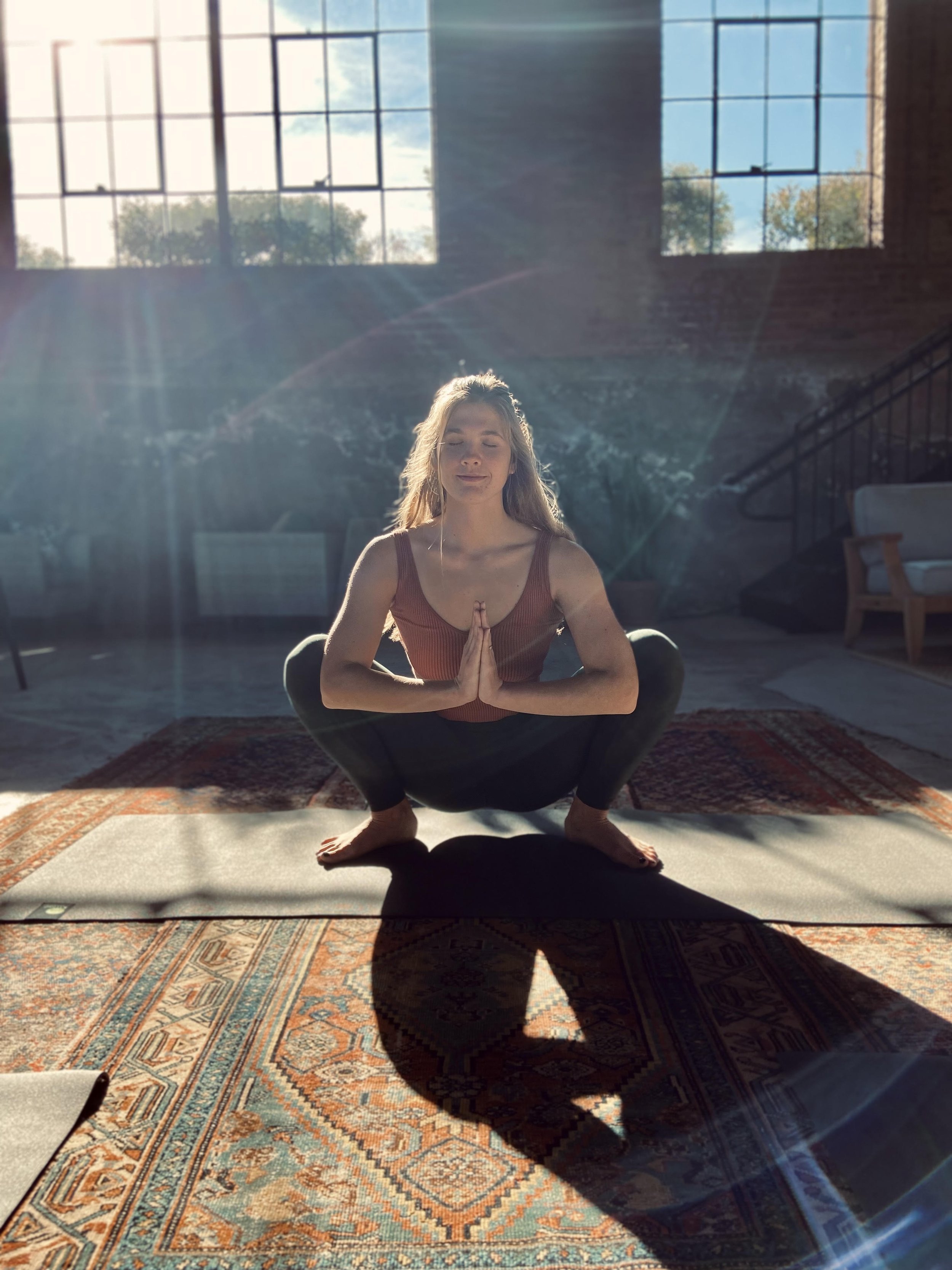 Joey-Lynn Henderson practicing yoga in a squatting pose on a mat, with sunlight streaming through large windows in a spacious room.