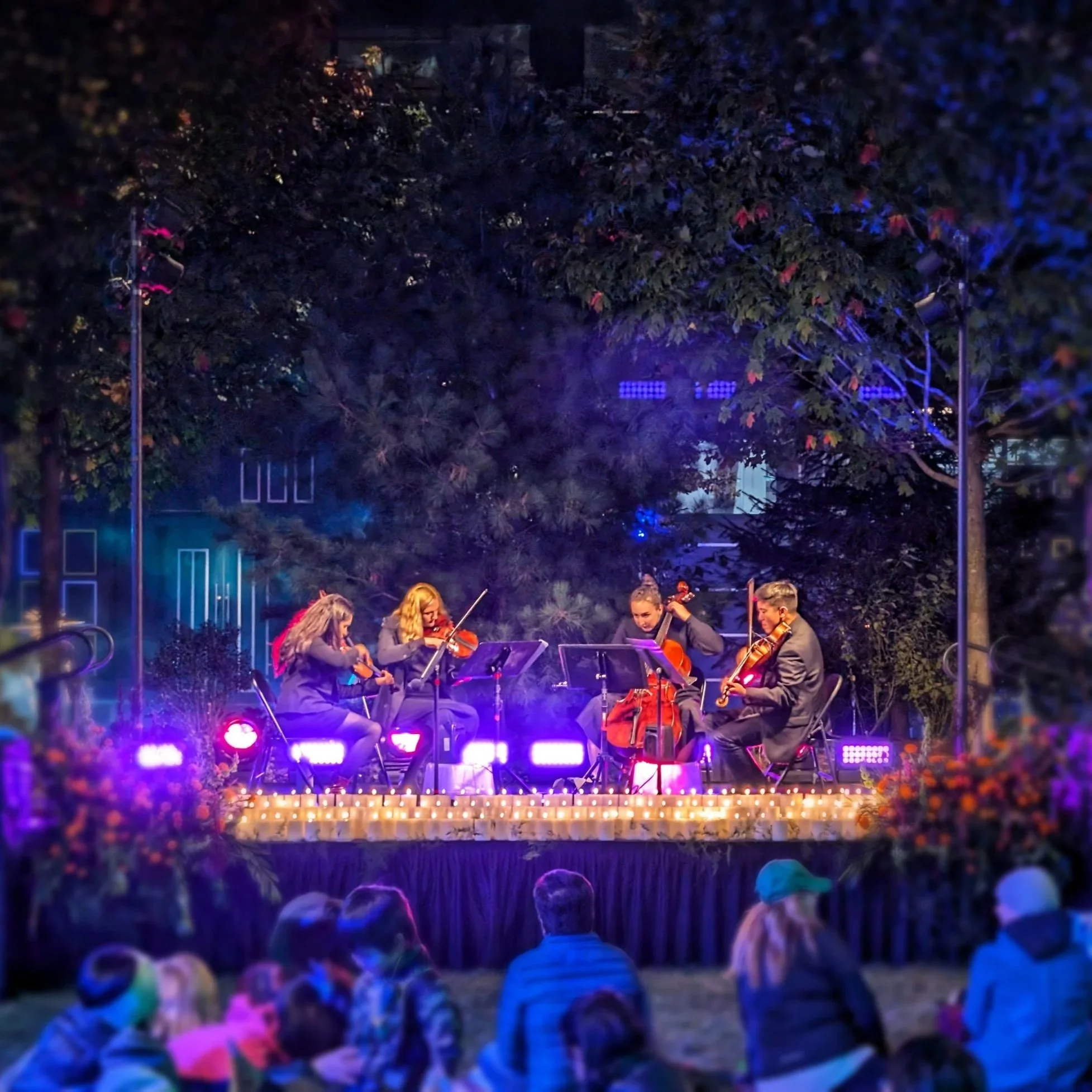 Night scene with a string quartet performing on an outdoor stage illuminated by purple and white lights, with audience members wearing colorful hats watching the performance. In the Seaport District, Boston, for their annual 'Play It By Fear' event.