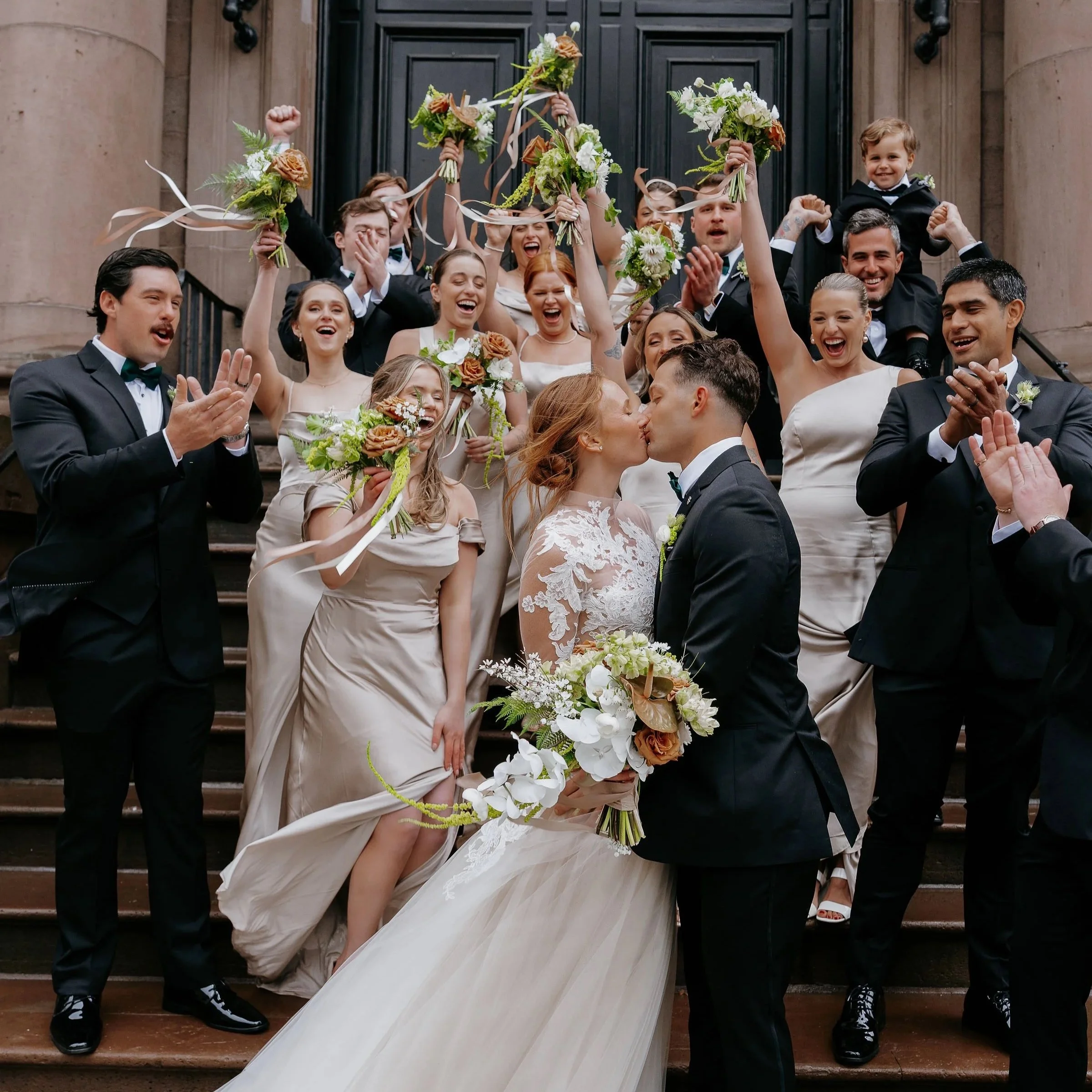 Group of people celebrating a wedding on the steps of a building, holding bouquets and kissing. Post recessional music.