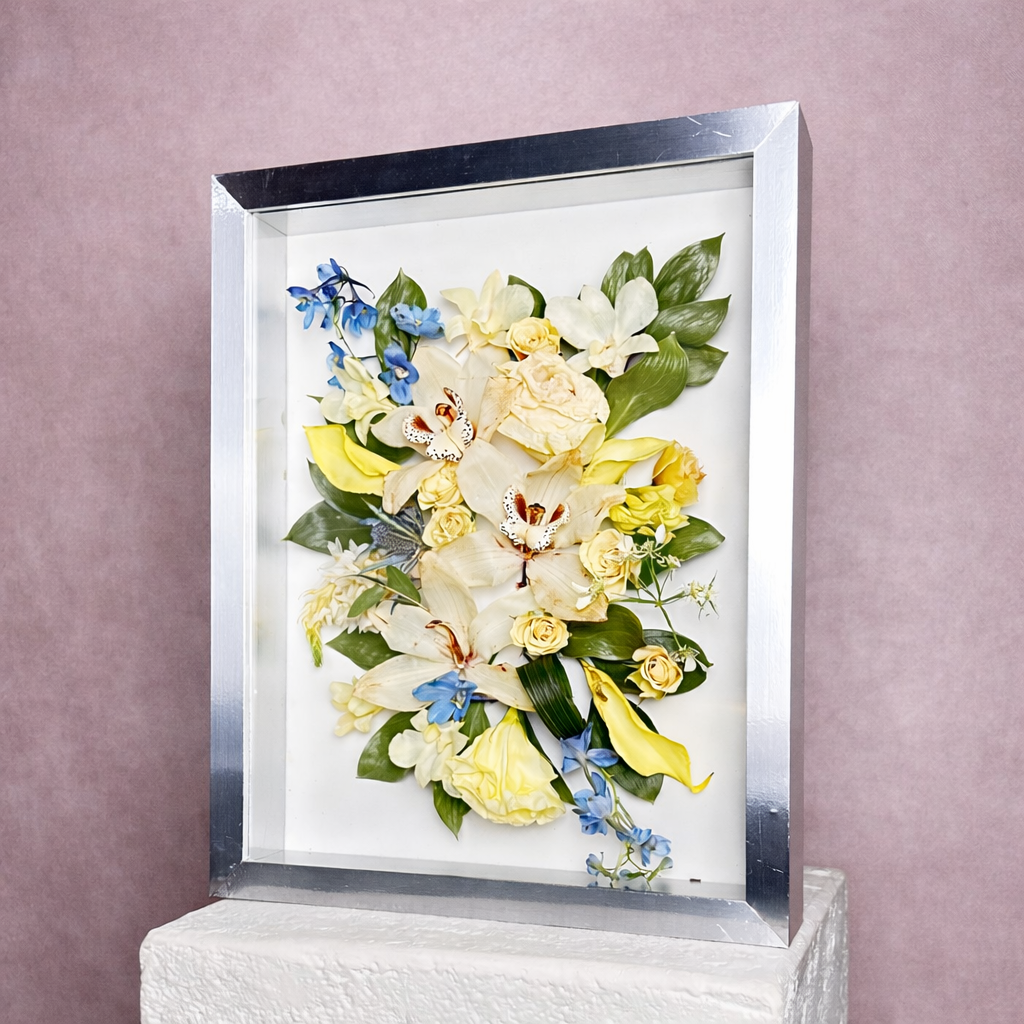 A framed 3D floral art piece with cream, yellow, blue, and green flowers and leaves, displayed on a wall.