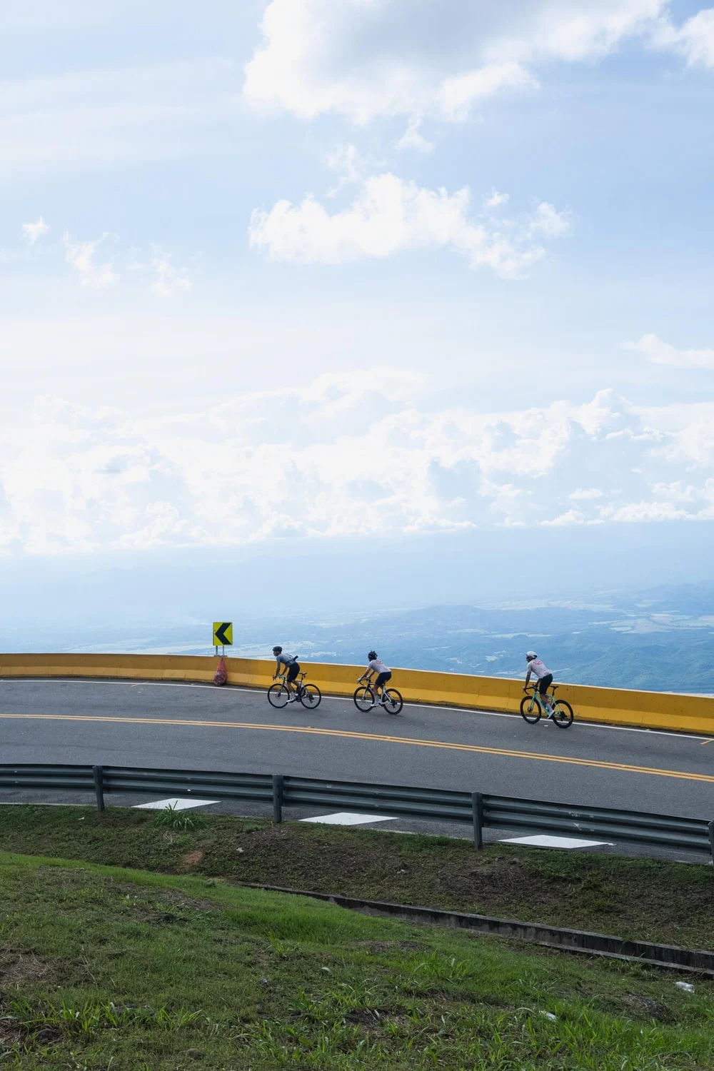 Three cyclists riding on a winding mountain road with a yellow guardrail, Into Coffee Heartlands