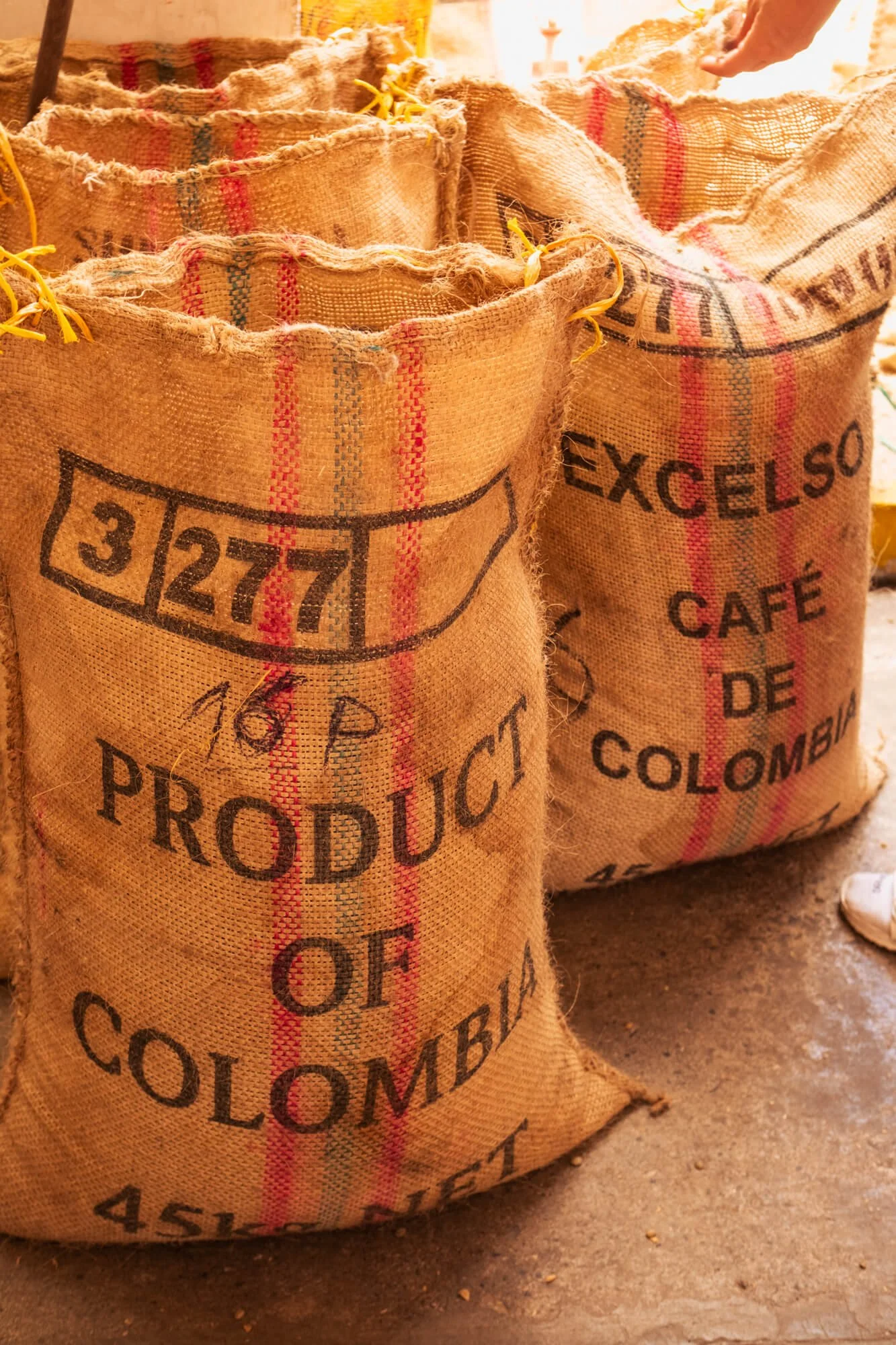 Bags of coffee with labels indicating they are product of Colombia, with one bag partially visible containing the text 'EXCELSO CAFÉ DE COLOMBIA' and another with the numbers '32771'.
