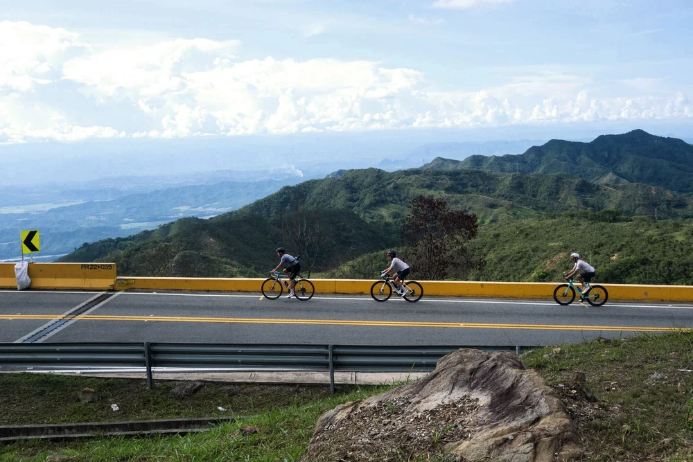 Three cyclists on a mountain road with green mountains and clouds