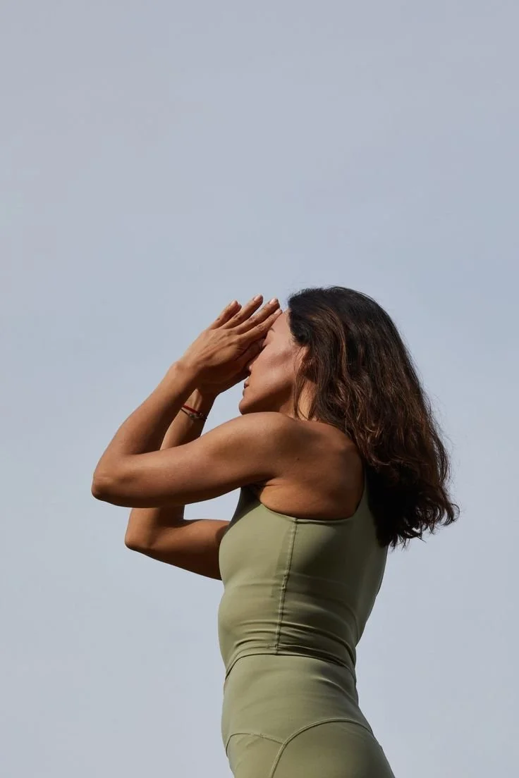 A woman practicing yoga outdoors with her hands in a prayer position near her face, wearing a green sports top, against a cloudy sky.