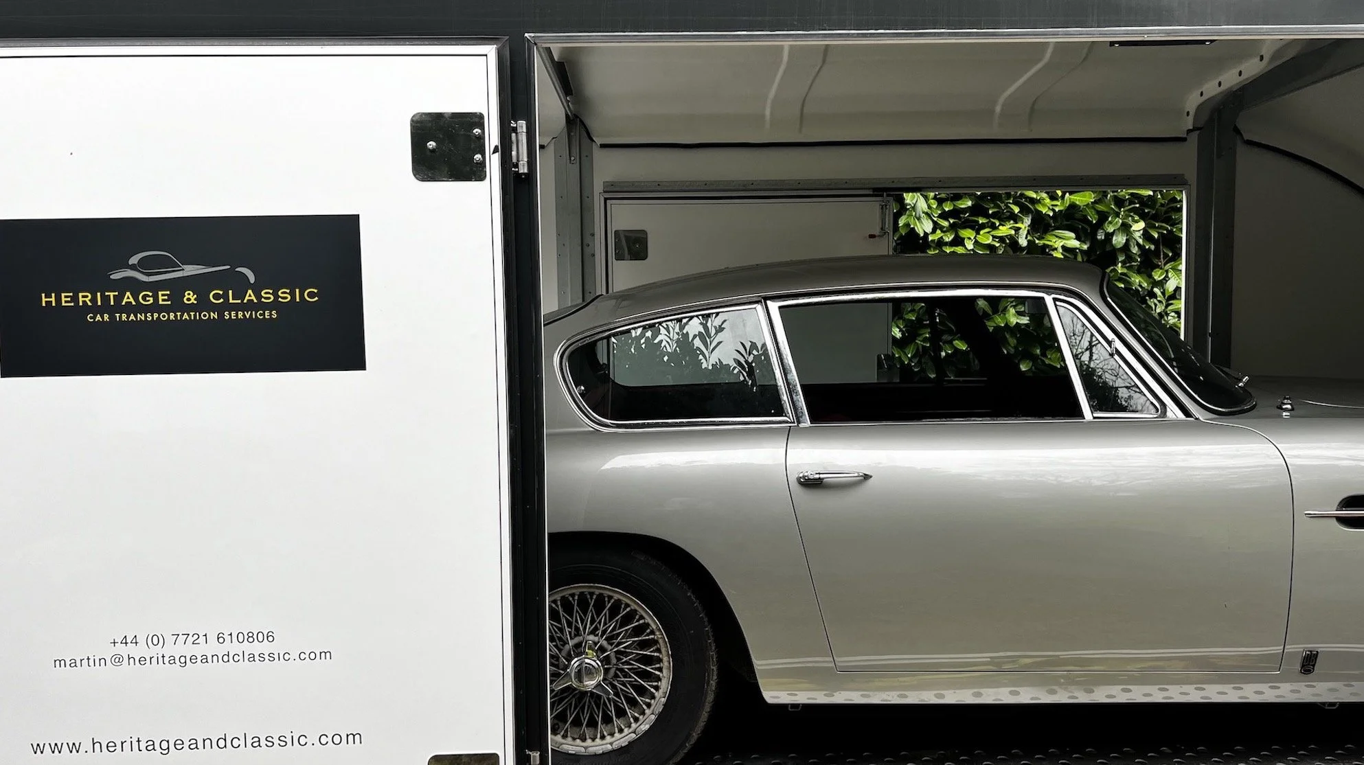 Silver Aston Martin DB6 inside a trailer with Heritage & Classic Car Transportation branding, offering services like vehicle inspection, tracking, and insurance.