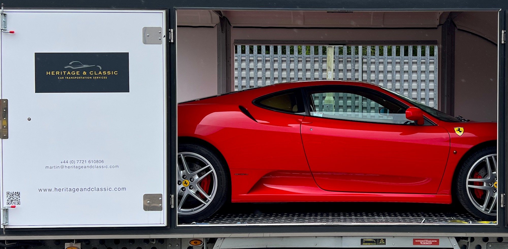 Red Ferrari inside a trailer with Heritage & Classic Car Transportation branding, offering services like vehicle inspection, tracking, and insurance.