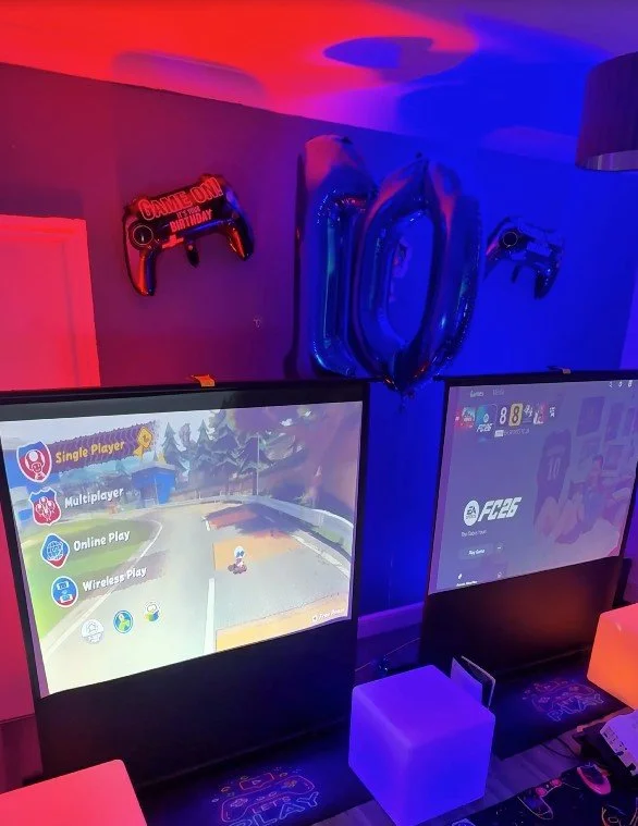 Two gaming monitors on a table illuminated with colorful LED lights. One screen shows a racing game menu with options like Single Player, Multiplayer, Online Play, and Wireless Play. Behind the monitors, there are large silver foil balloons spelling 