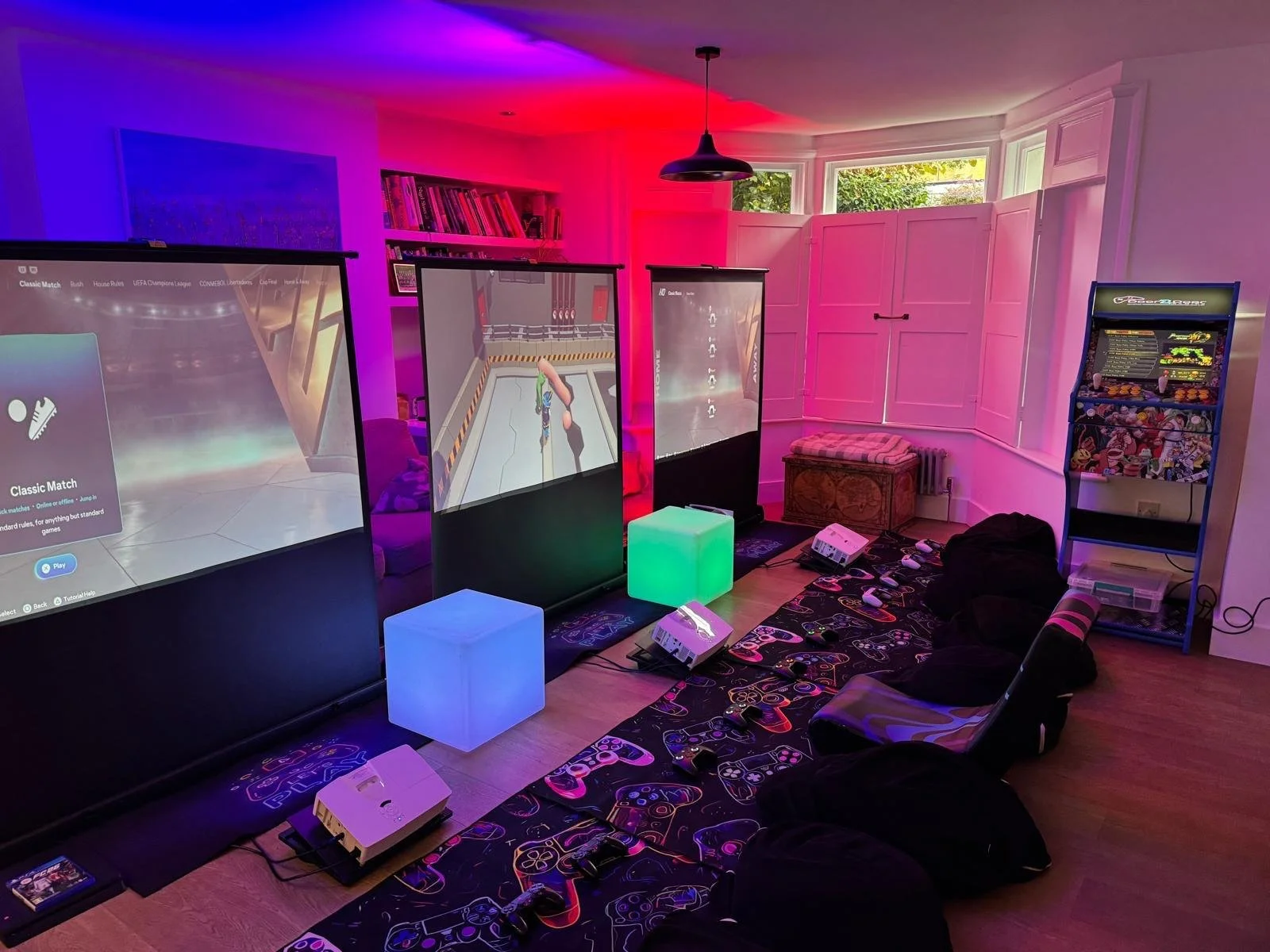 A gaming room with three large screens displaying video games, illuminated by colorful LED lights, with gaming consoles, controllers, a pinball machine, and lagging bags on the floor.