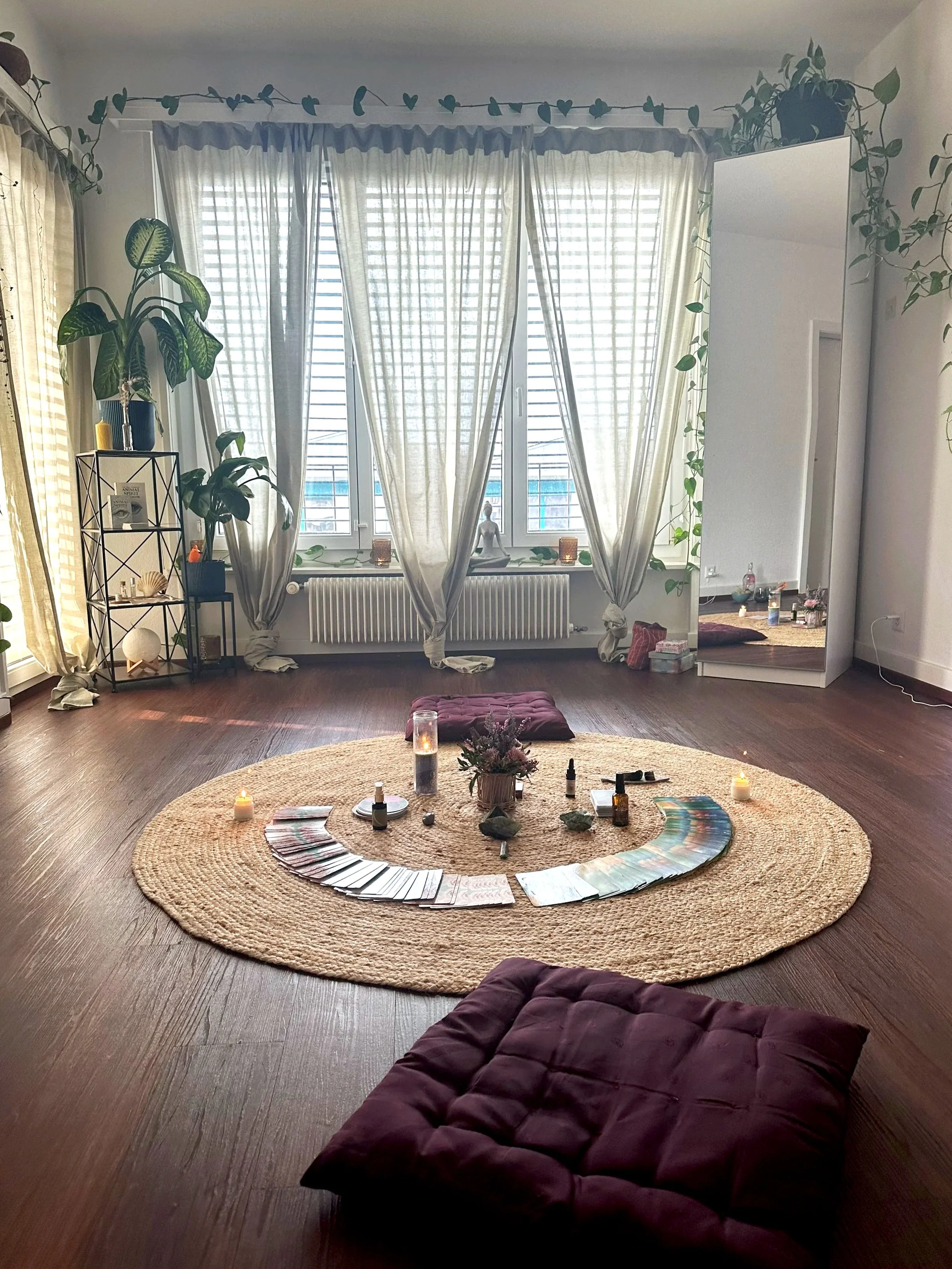 Une pièce calme et lumineuse avec des rideaux blancs, une moquette ronde, des plantes vertes, des bougies allumées, des cristaux, une bougie gel, des huiles essentielles, et des tapis de méditation, créant une ambiance zen pour la méditation ou la relaxation.