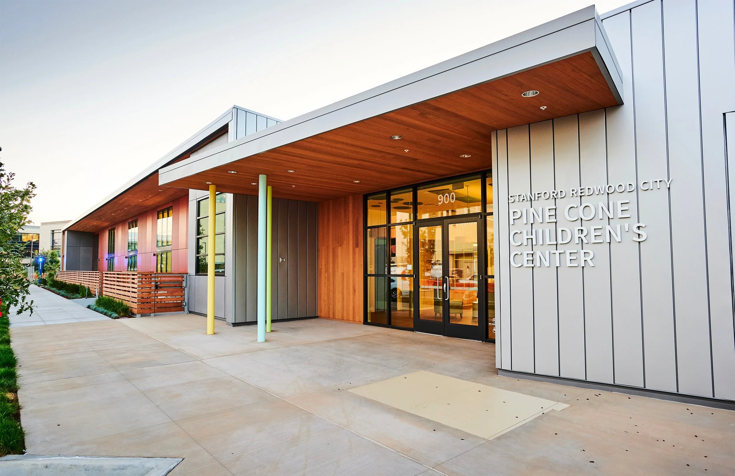 Exterior of the Pine Cone Children's Center at Stanford Redwood City, showing modern building with wood accents, colorful poles, and glass doors.