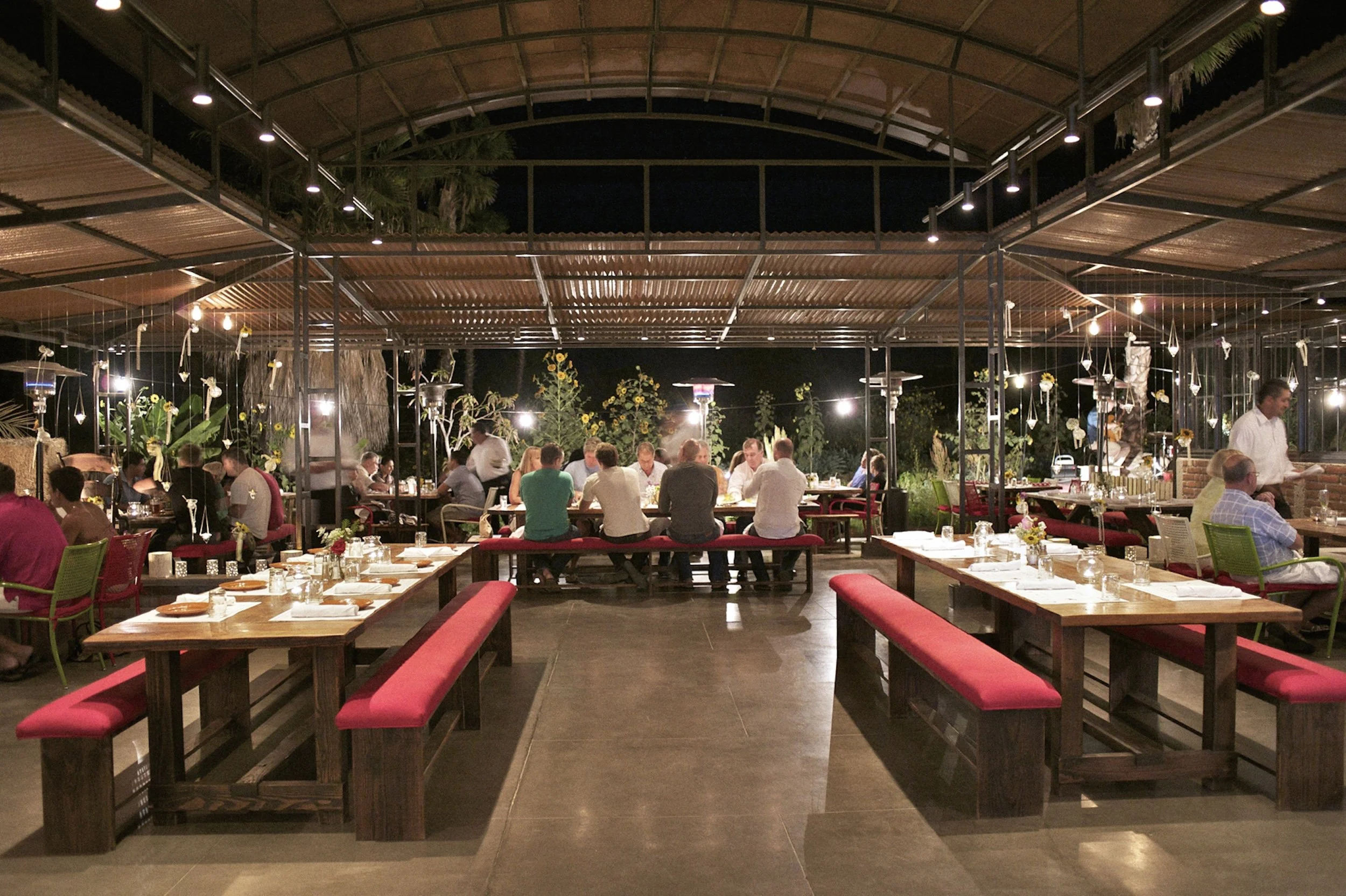 A lively restaurant at night with people seated at tables, warm lighting, and outdoor plants.