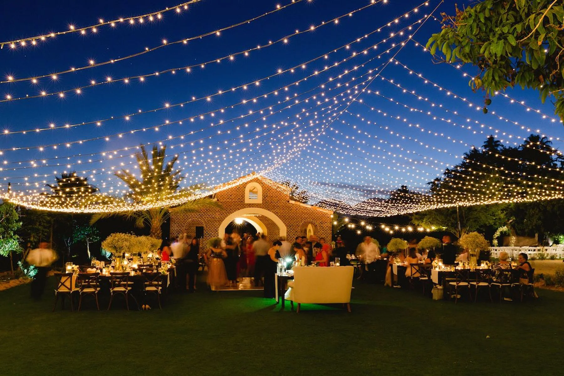 Outdoor evening event with string lights overhead, guests mingling and dining around tables, and a brick building in the background.