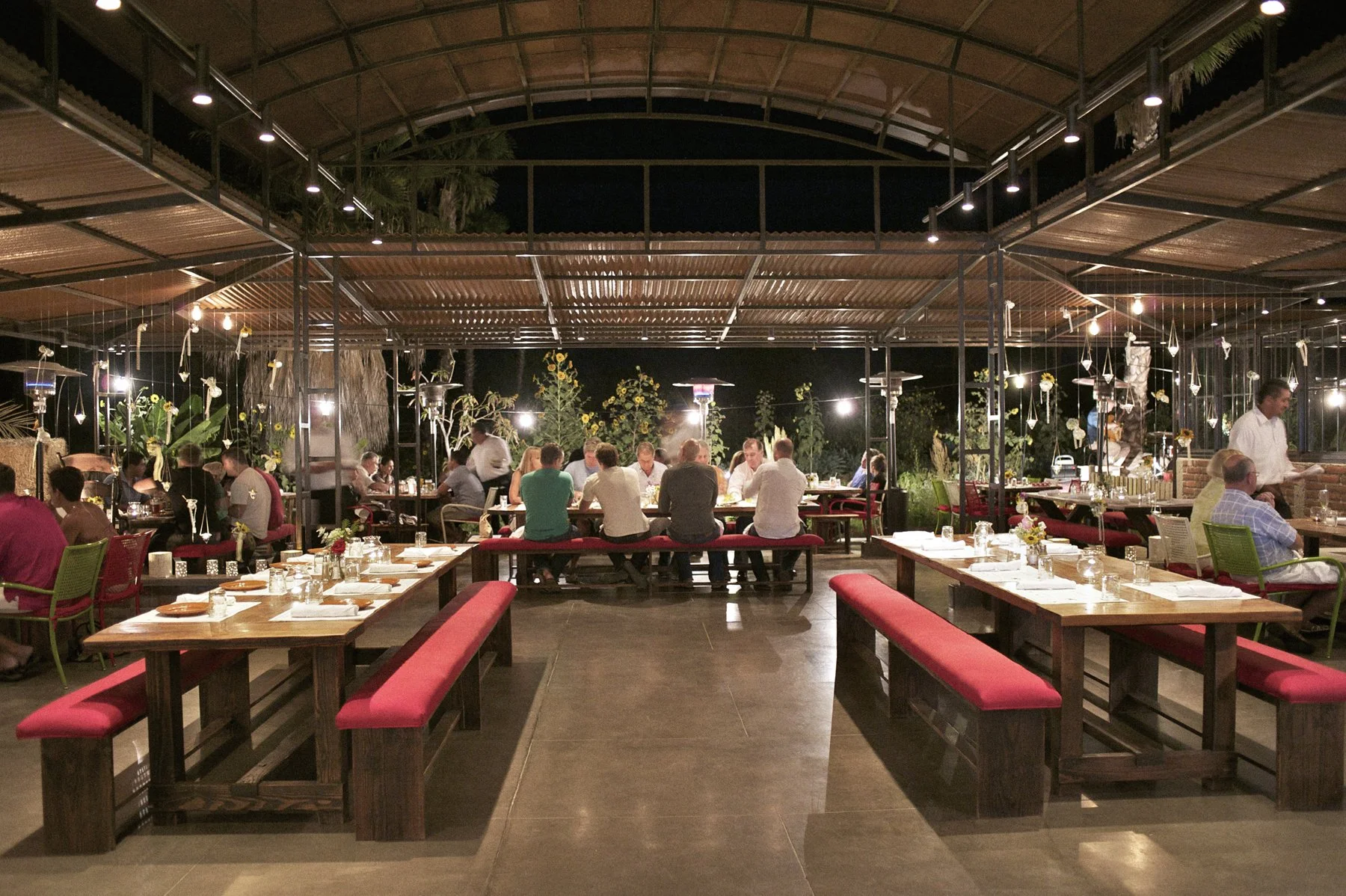 Outdoor restaurant at night with tables, red benches, and diners, illuminated by string lights and heaters, under a high, curved wooden roof.