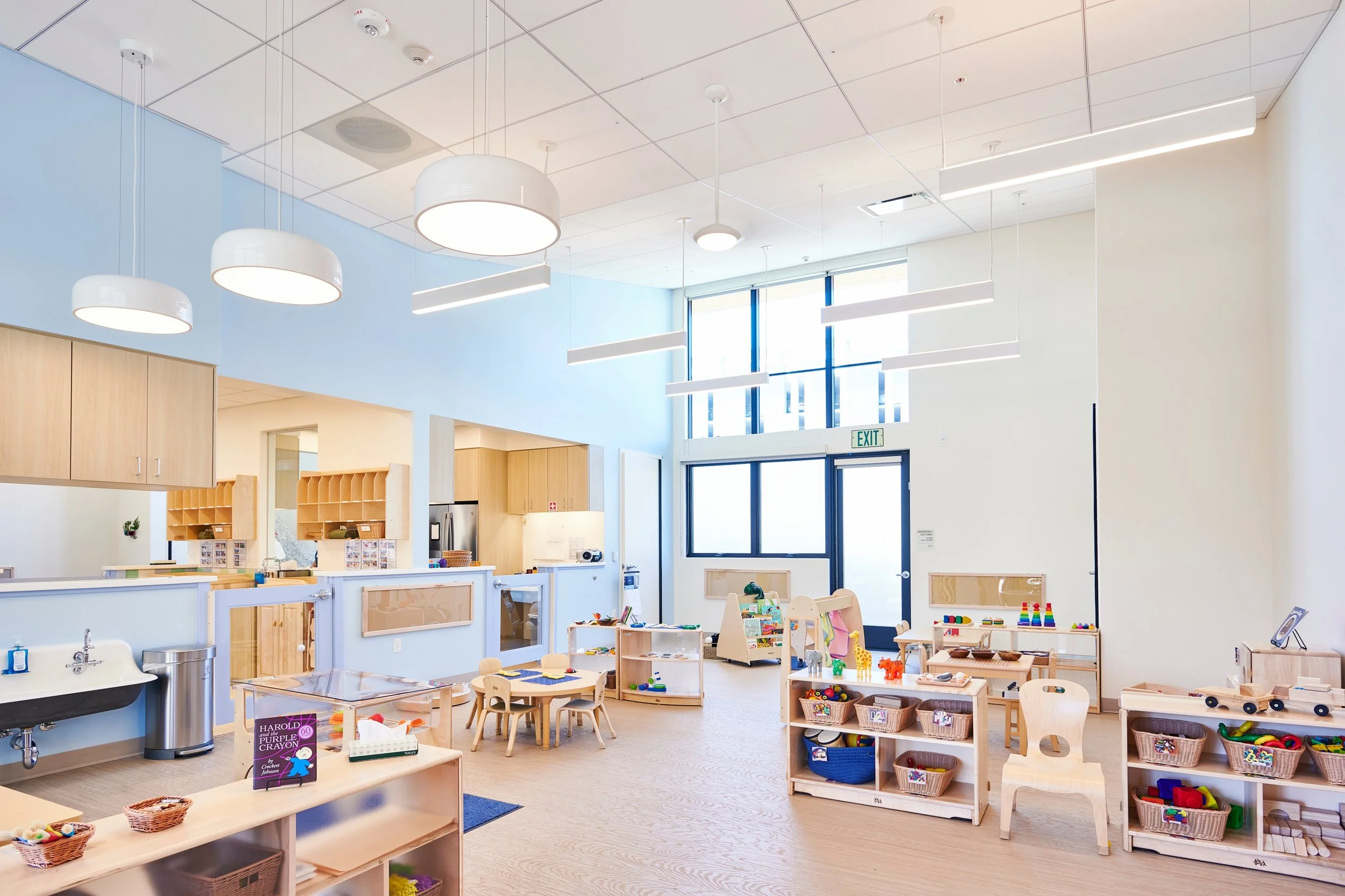 Bright, well-lit classroom with light wood furniture, toys, and educational materials for young children, large front windows, and modern ceiling lights.