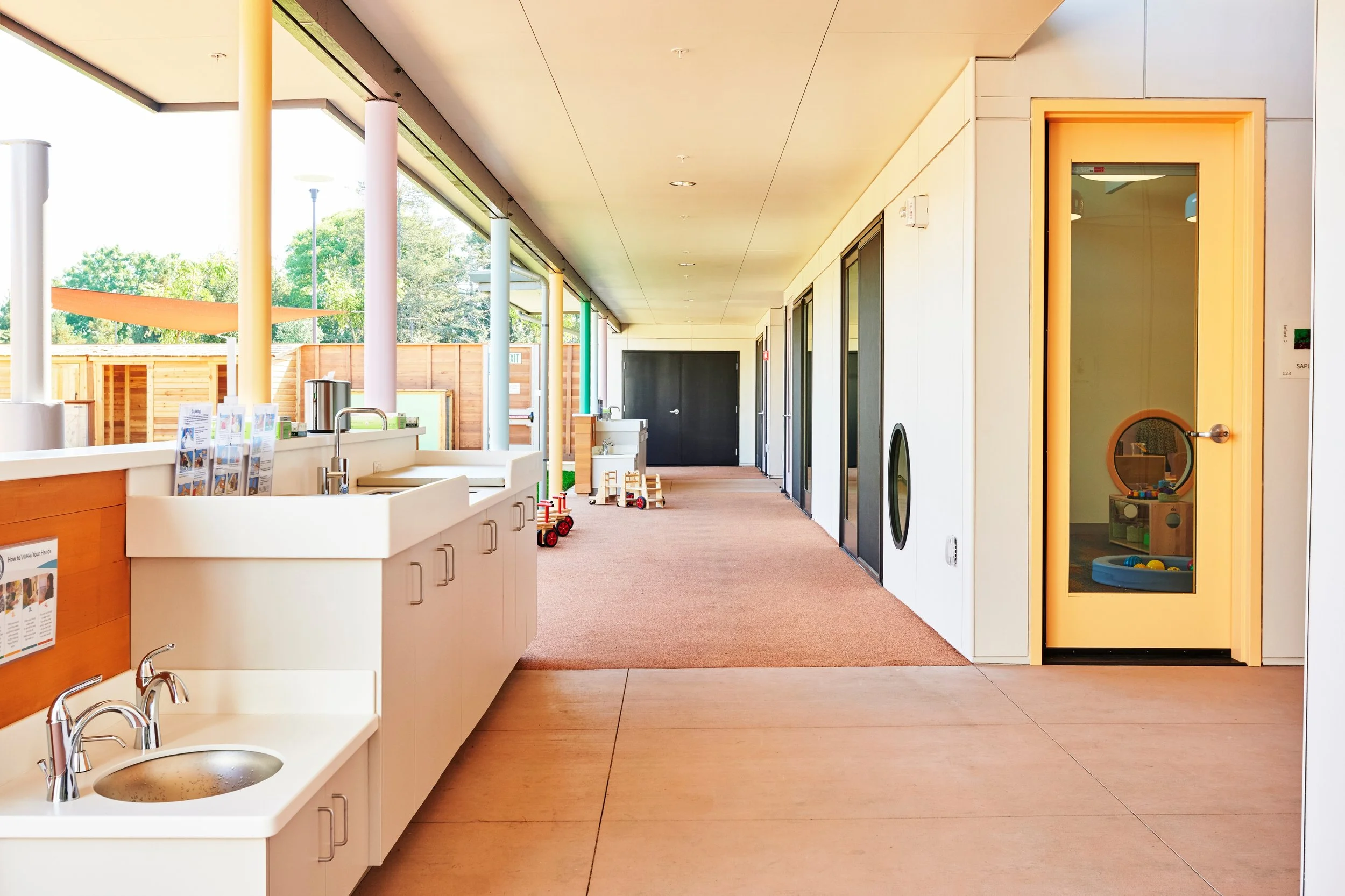 Children's indoor play area with small sinks, child's door with window, and toys, illuminated by natural light from large windows.