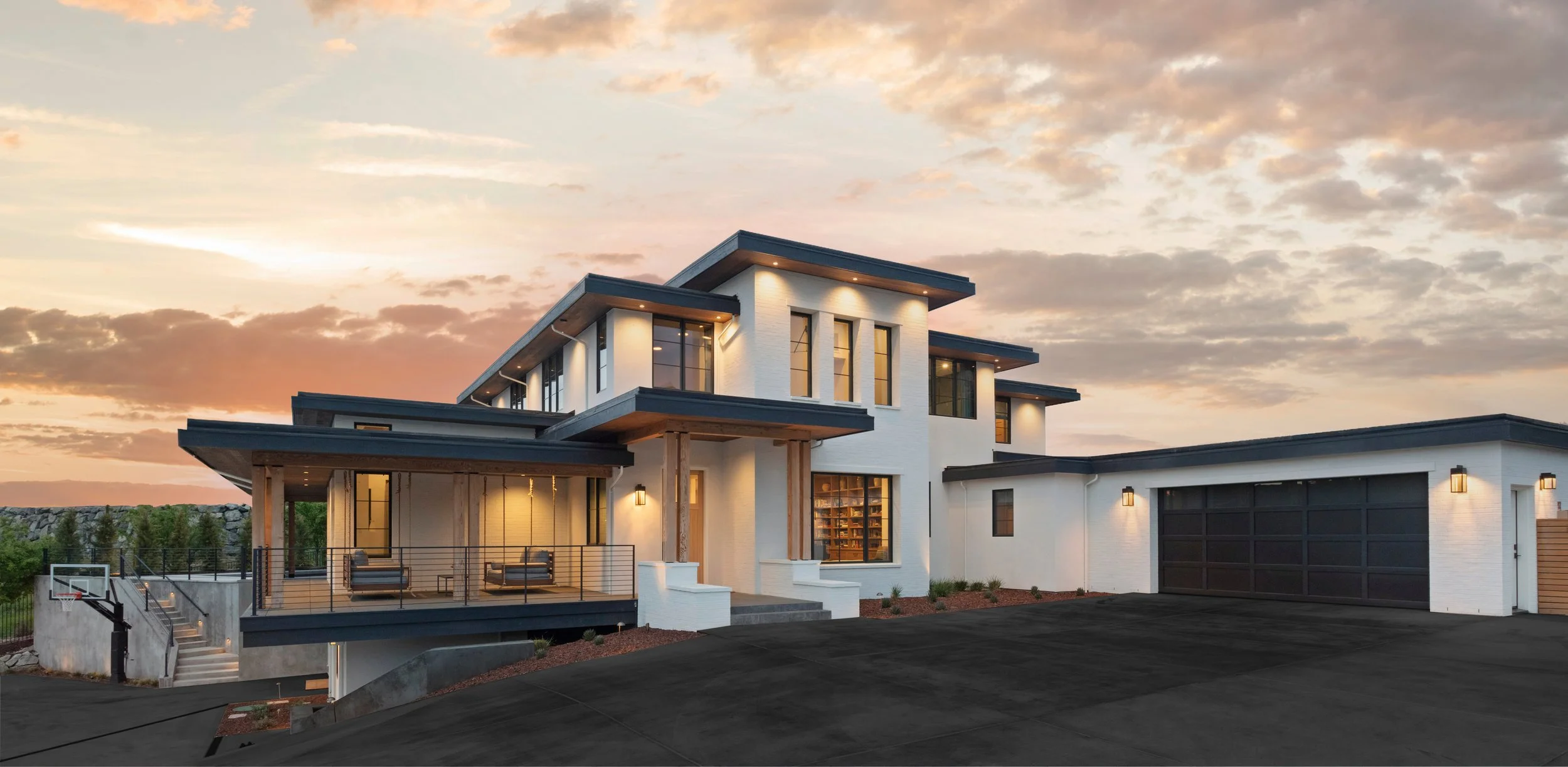 Modern white multi-story house with a large garage, illuminated exterior lights, a porch with outdoor seating, and a sunset sky in the background.