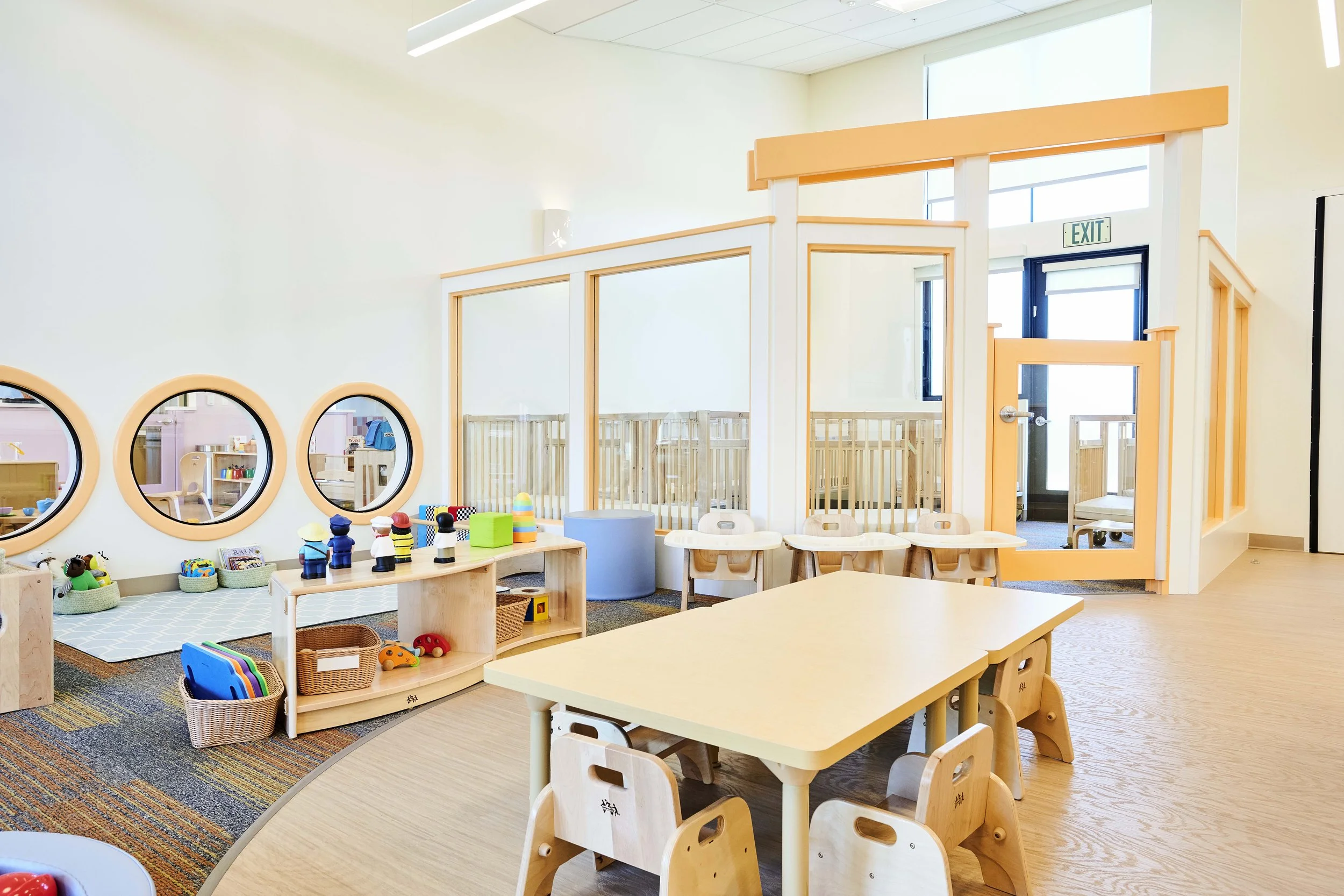 Bright preschool classroom with wooden tables and chairs, colorful toys, circular windows, and a child-proof enclosed play area.