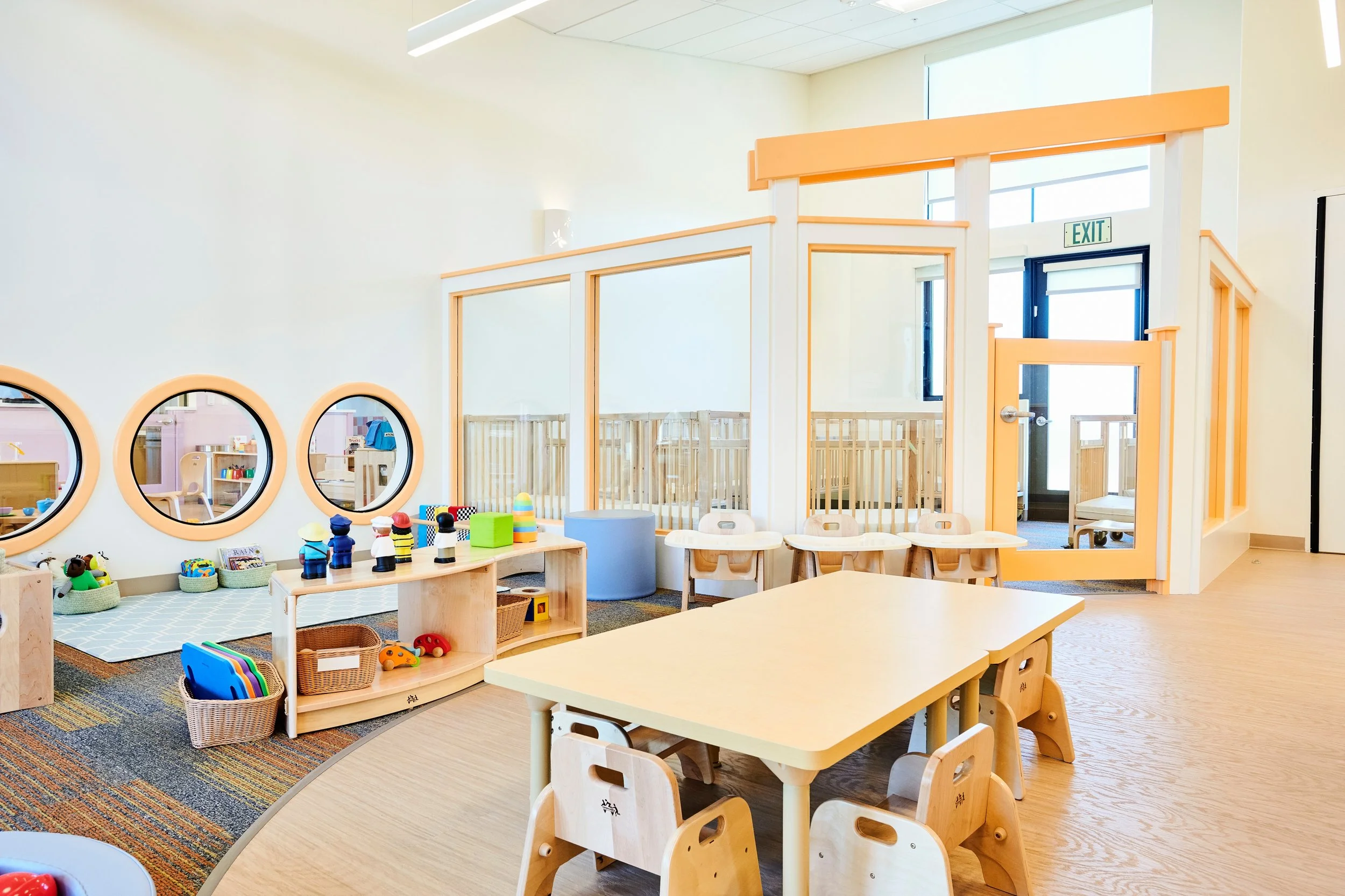 Bright, modern children's playroom with small tables and chairs, colorful toys, and a nursery area separated by a glass partition. Windows allow natural light to fill the space.