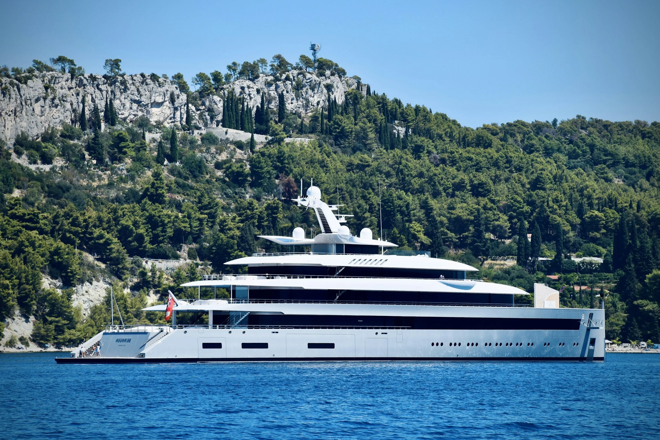 Large white modern yacht on blue water with green forested hillside in the background.