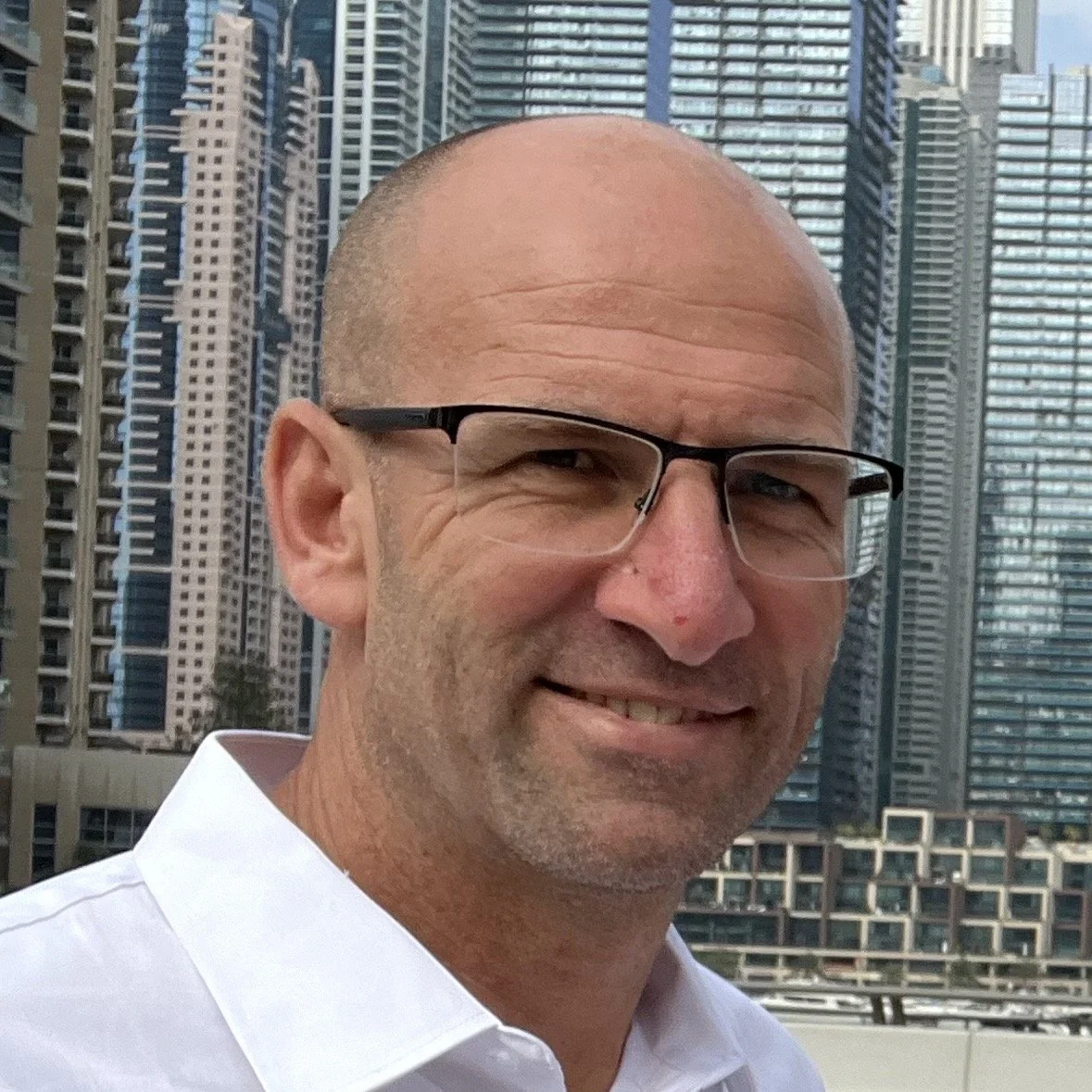 A middle-aged man with glasses, a bald head, and a beard, smiling in front of a cityscape with tall skyscrapers.