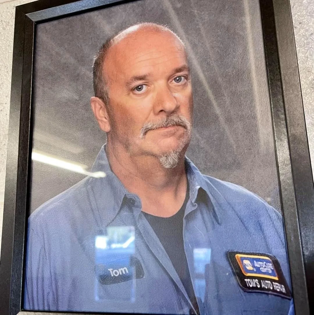 Framed portrait of a middle-aged man with a mustache and goatee, wearing a blue work uniform with a name tag that reads 'Tom' and a patch indicating he is an auto technician.