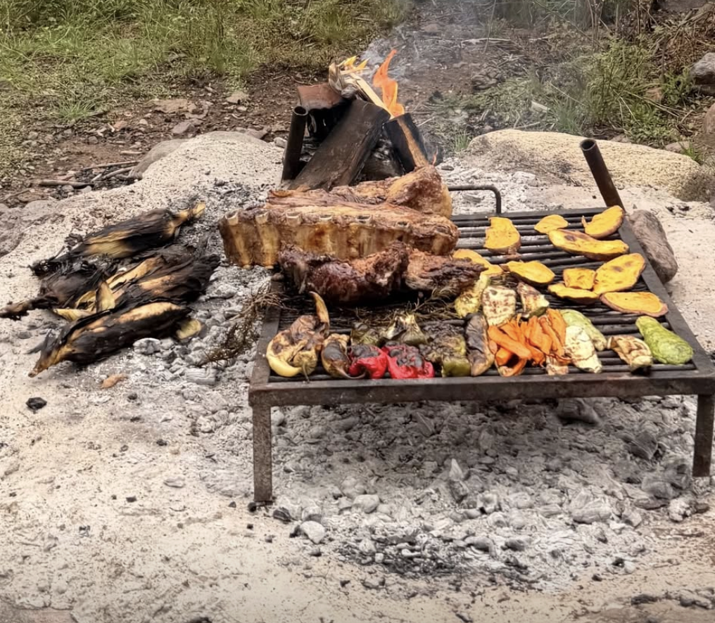 Outdoor barbecue grill with cooked food including roasted corn, grilled vegetables, and a large piece of meat over an open fire.