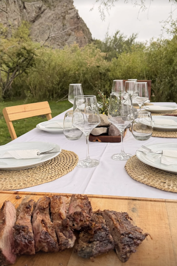 Outdoor dining table set with plates, glasses, and a centerpiece of rocks and greenery, with a background of trees and rocky hillside, and a cutting board with cooked meat in the foreground.