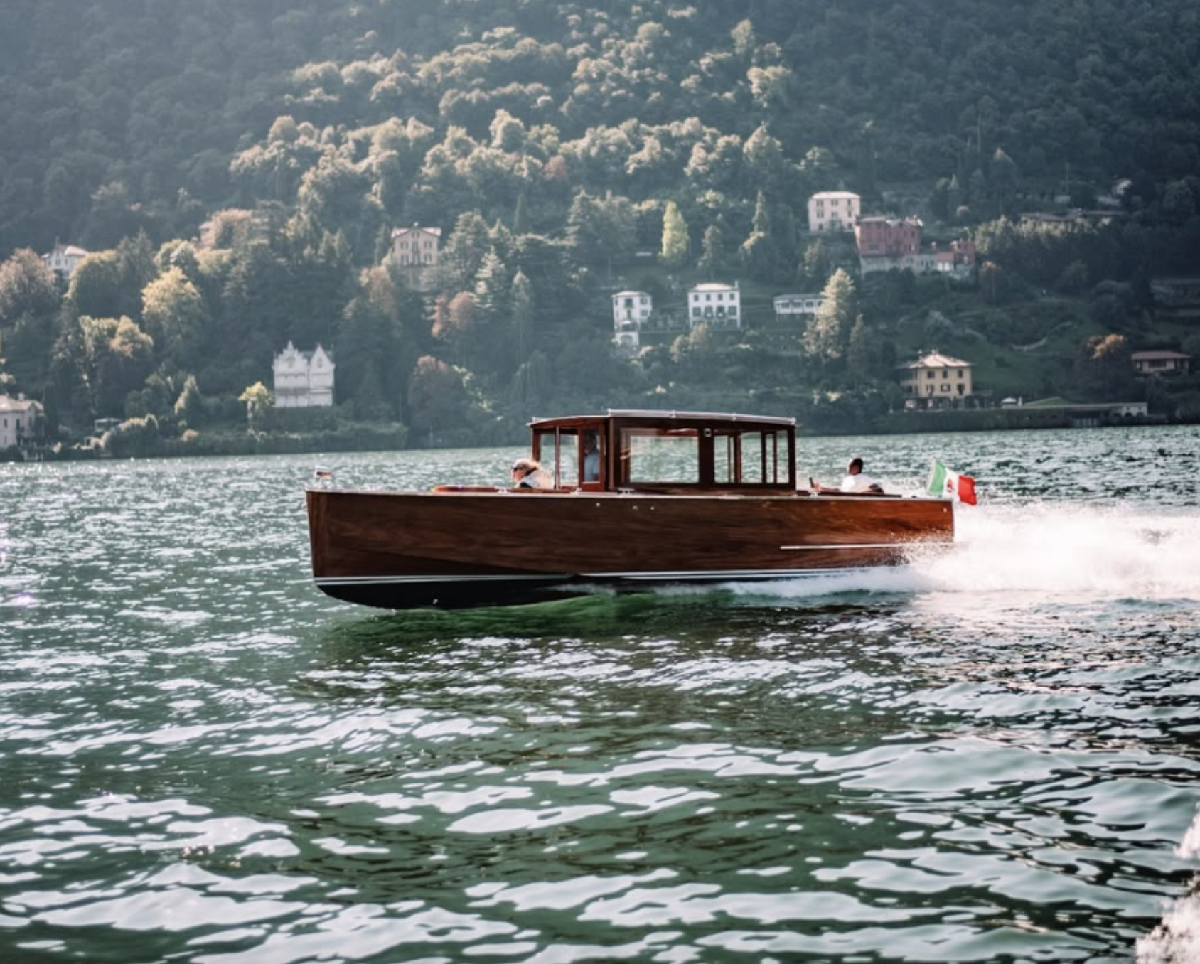 A wooden motorboat speeds across a body of water with a scenic hillside town in the background, featuring houses and trees.
