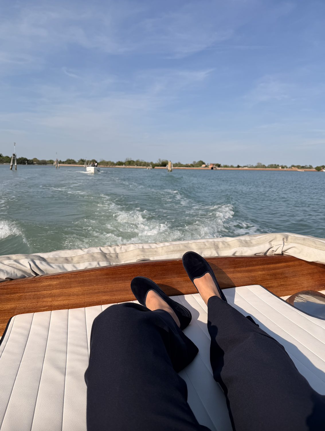 Person relaxing on a boat with black shoes and dark pants, view of water and sky, with other boats in the distance and shoreline