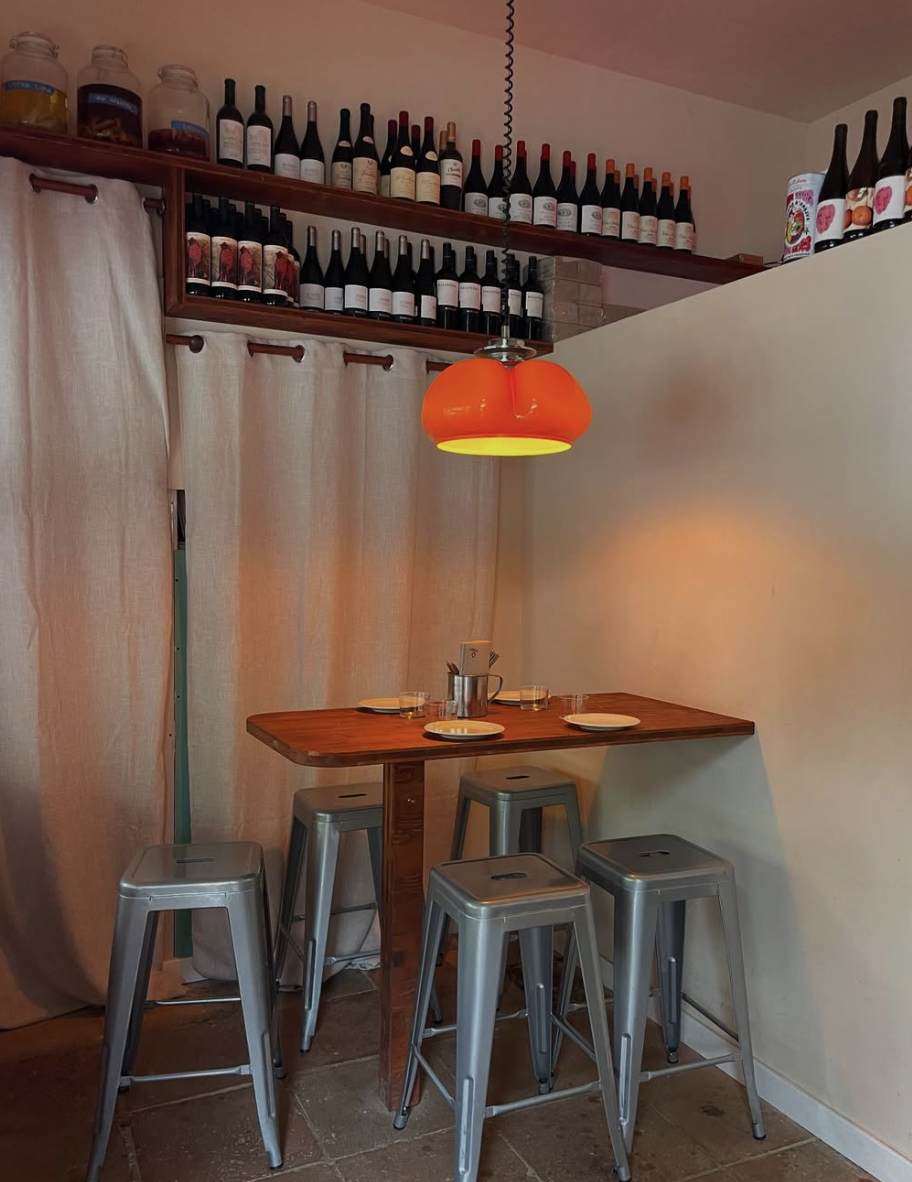 A cozy dining corner with a high wooden table set for four, surrounded by five metallic stools. An orange pendant light hangs above the table, casting warm light. The back wall features beige curtains covering a window, with shelves above displaying rows of wine bottles and decorative jars.