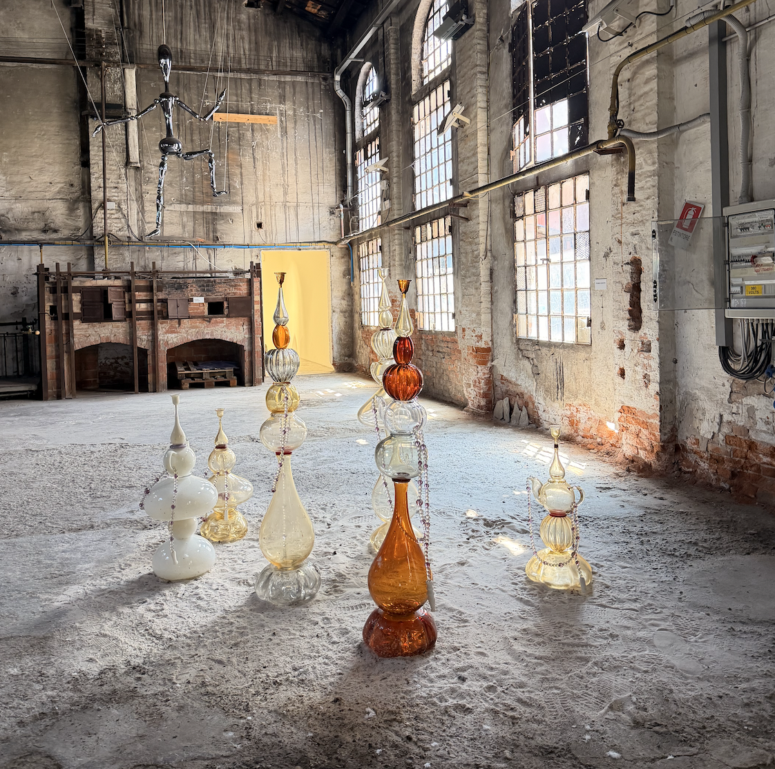 An art installation featuring tall, colorful glass sculptures inside an industrial warehouse with large windows, exposed brick walls, and a metal sculpture of a humanoid figure hanging from the ceiling.