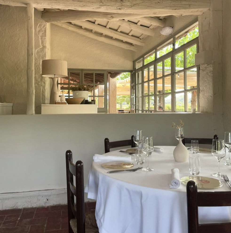 A round dining table set for a meal with white tablecloth, plates, wine glasses, water glasses, and a white vase with flowers in a bright restaurant with large windows and natural light.