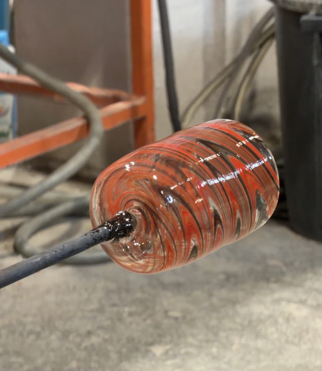 A close-up of a colorful, swirled glass object on a metal rod in a workshop setting.