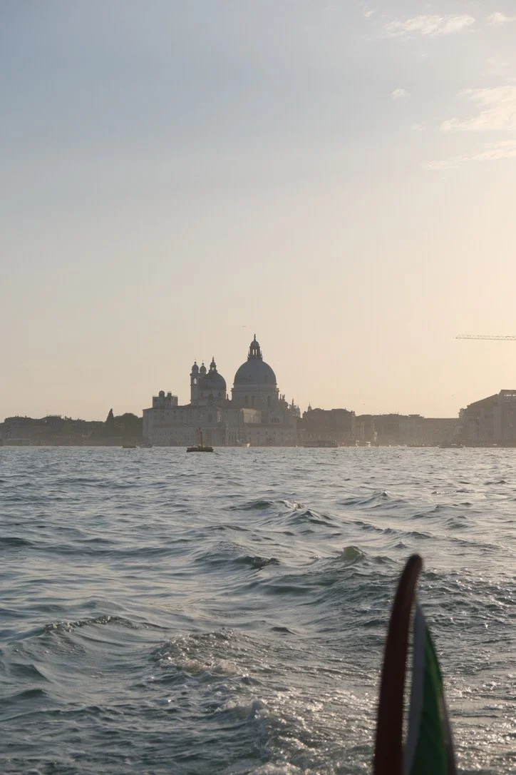 View of Venice, Italy, from the water, with water waves in the foreground and historic buildings and a domed church in the background at sunset.