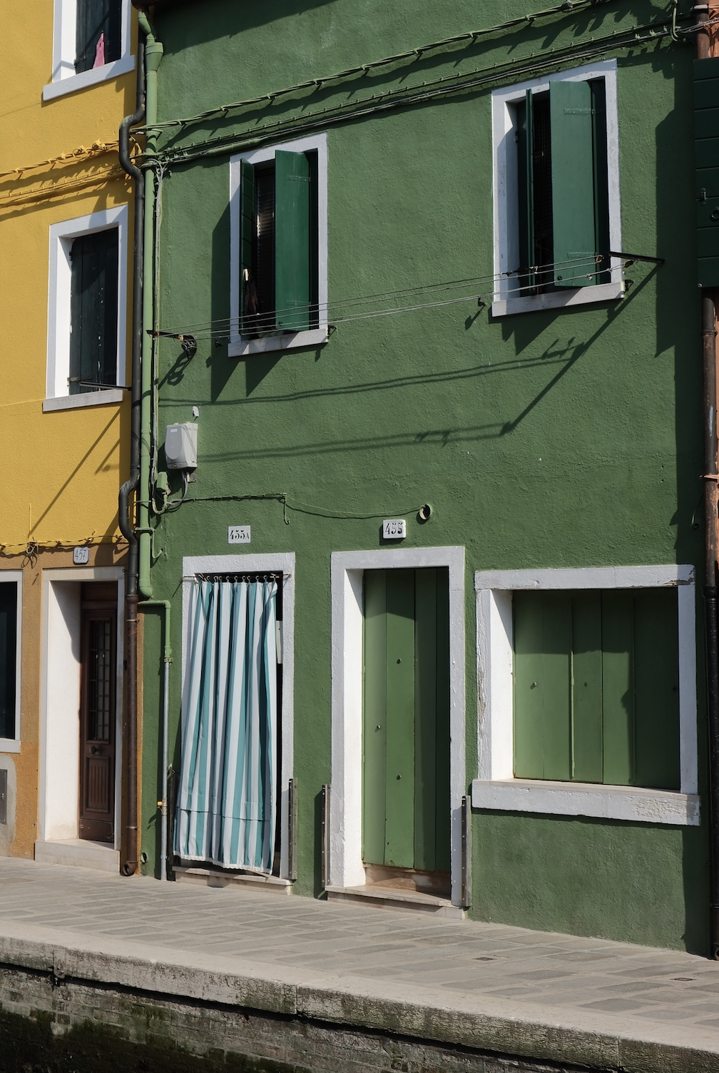 Colorful buildings in Italy, green and yellow, with windows and a door with a striped curtain, along a canal walkway.