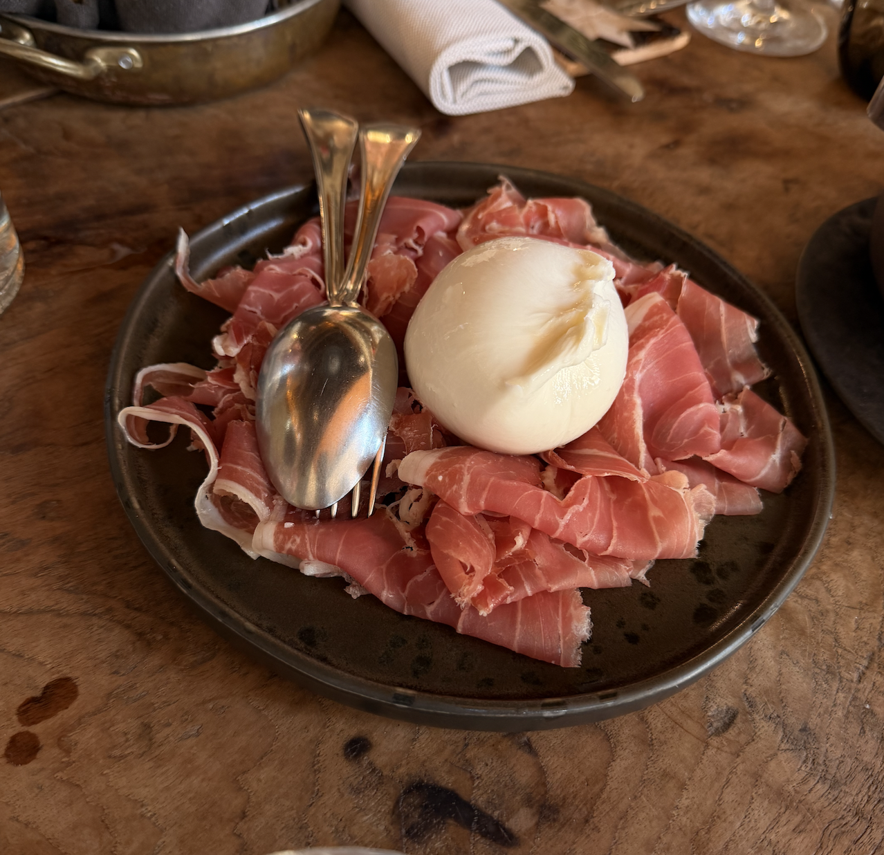 A black plate with sliced prosciutto and a ball of fresh mozzarella cheese, accompanied by a spoon and a fork, on a wooden table.