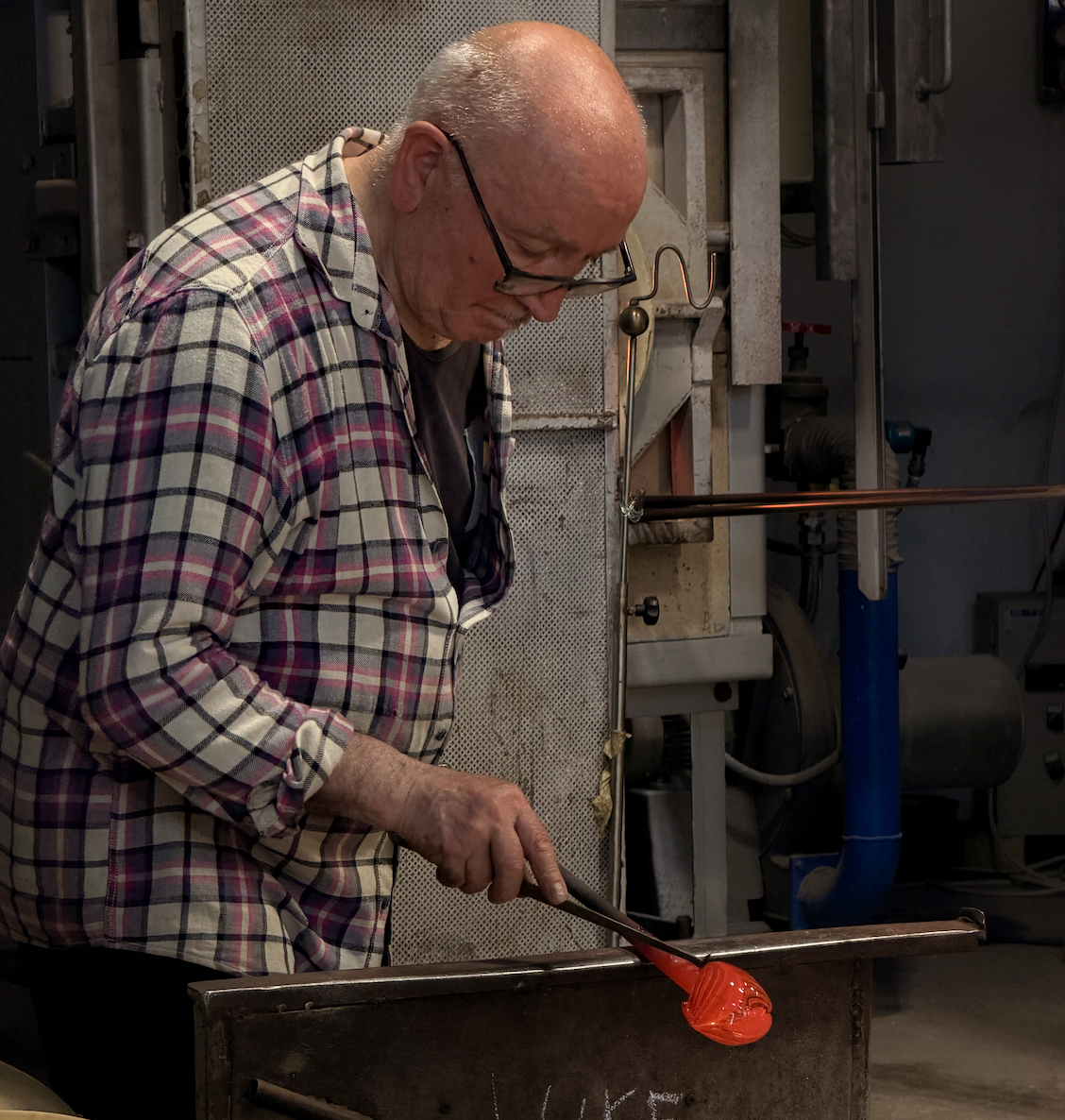 A man with glasses and a plaid shirt working on glassblowing in a workshop, shaping a glowing orange molten piece with a tool.